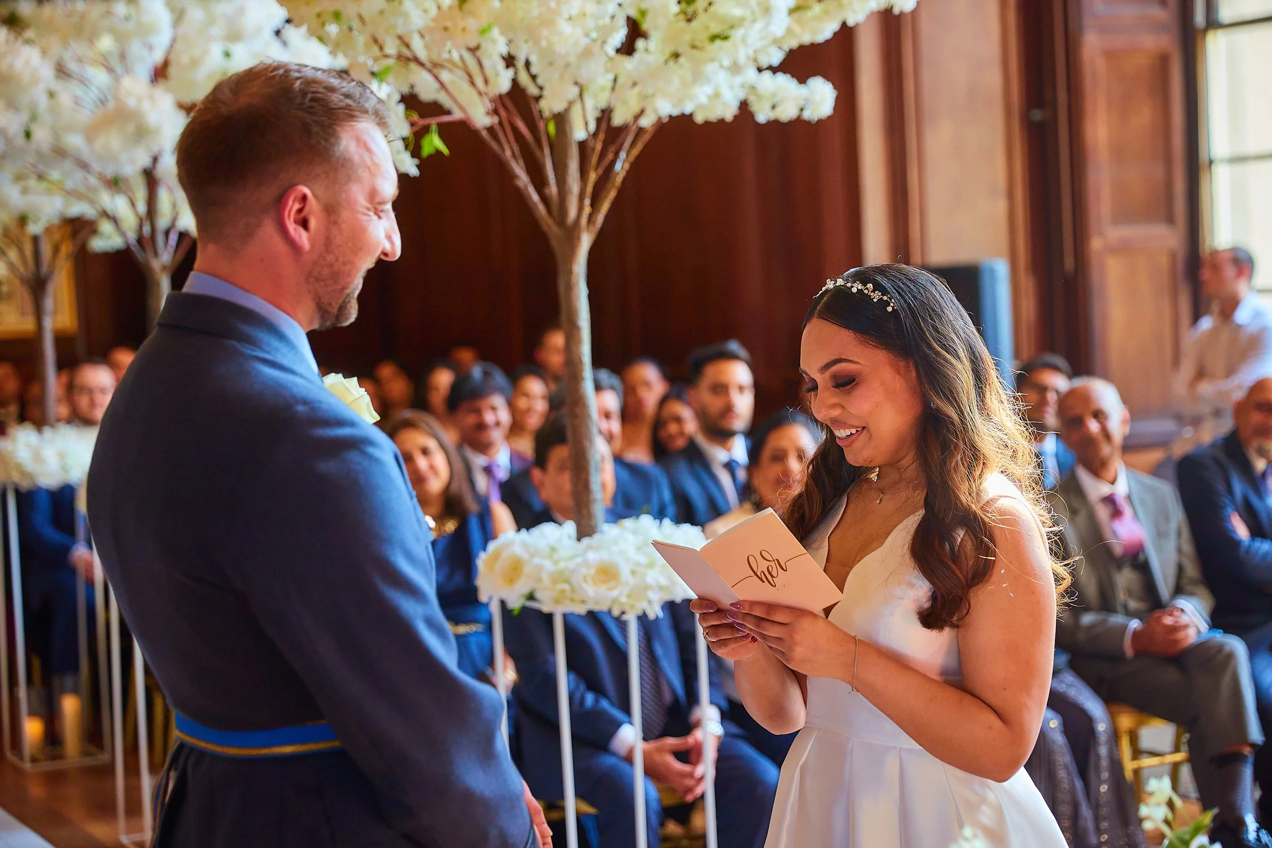 Bride and groom exchanging vows during wedding ceremony in elegant indoor setting, with guests seated in the background.
