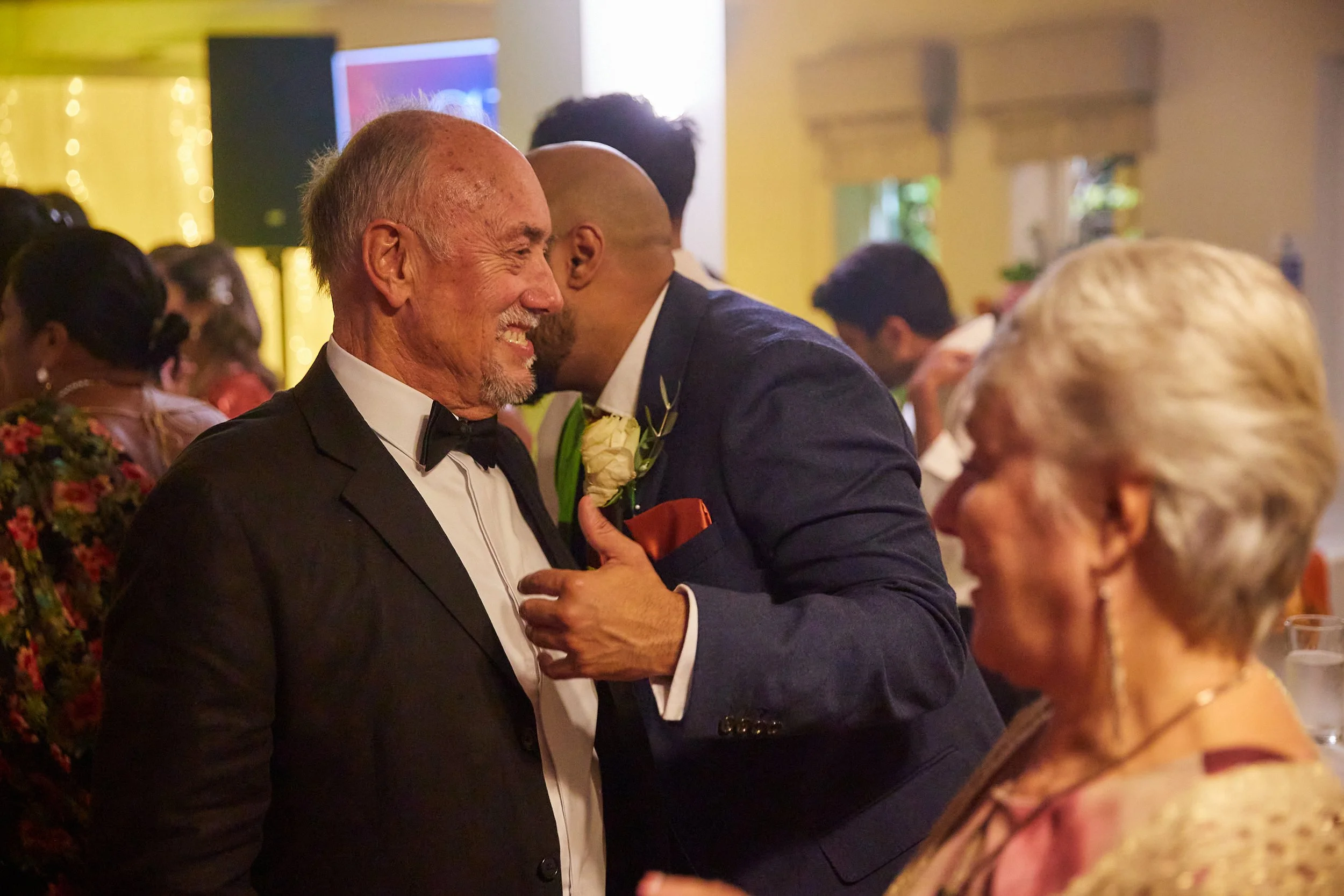 A joyful elderly man in a tuxedo and bow tie smiling at a man in a suit with a boutonniere, while a woman with white hair and a gold dress looks on at a formal event or celebration.