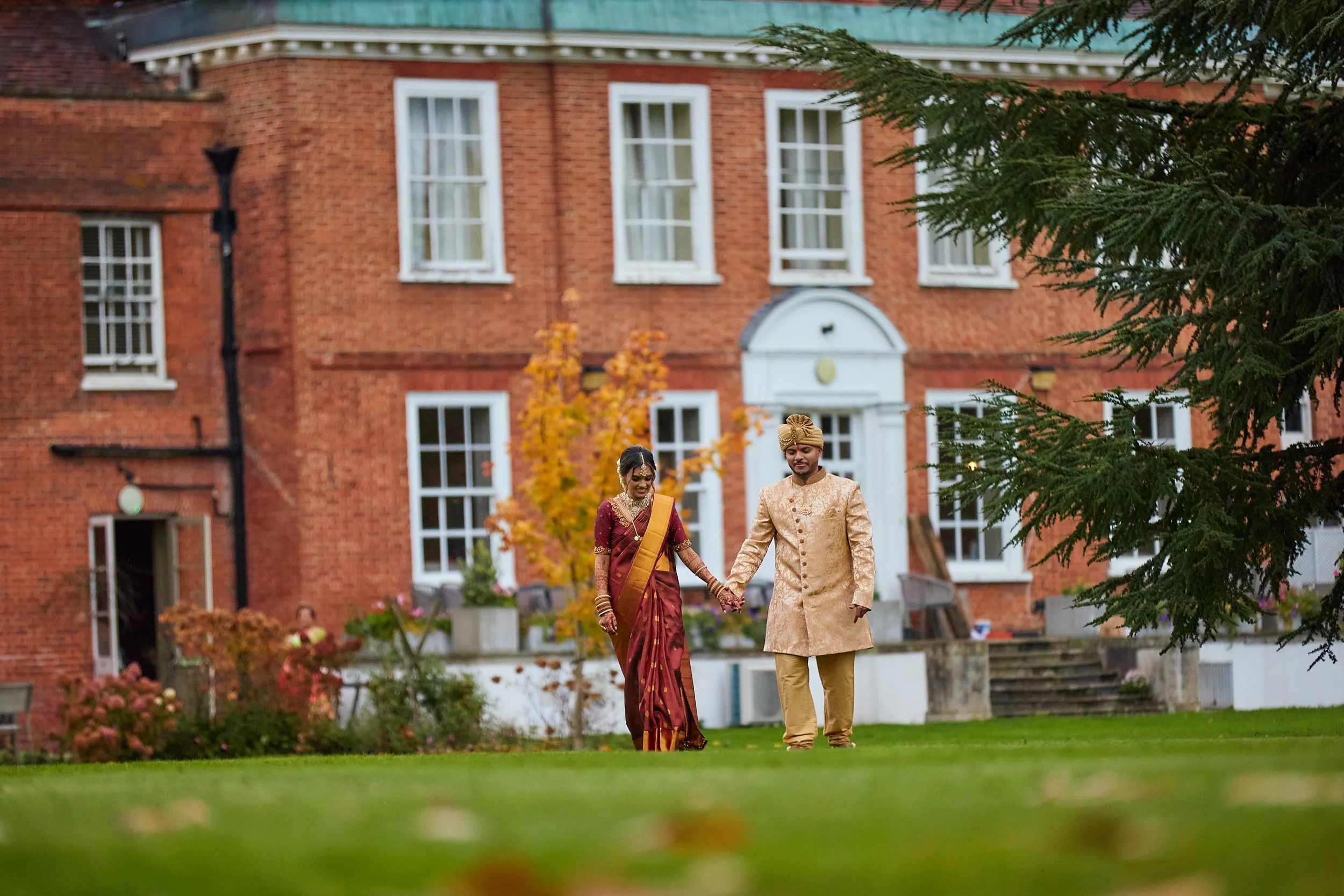 A newlywed Indian couple dressed in traditional wedding attire walking hand in hand on a lawn in front of a large red brick house.