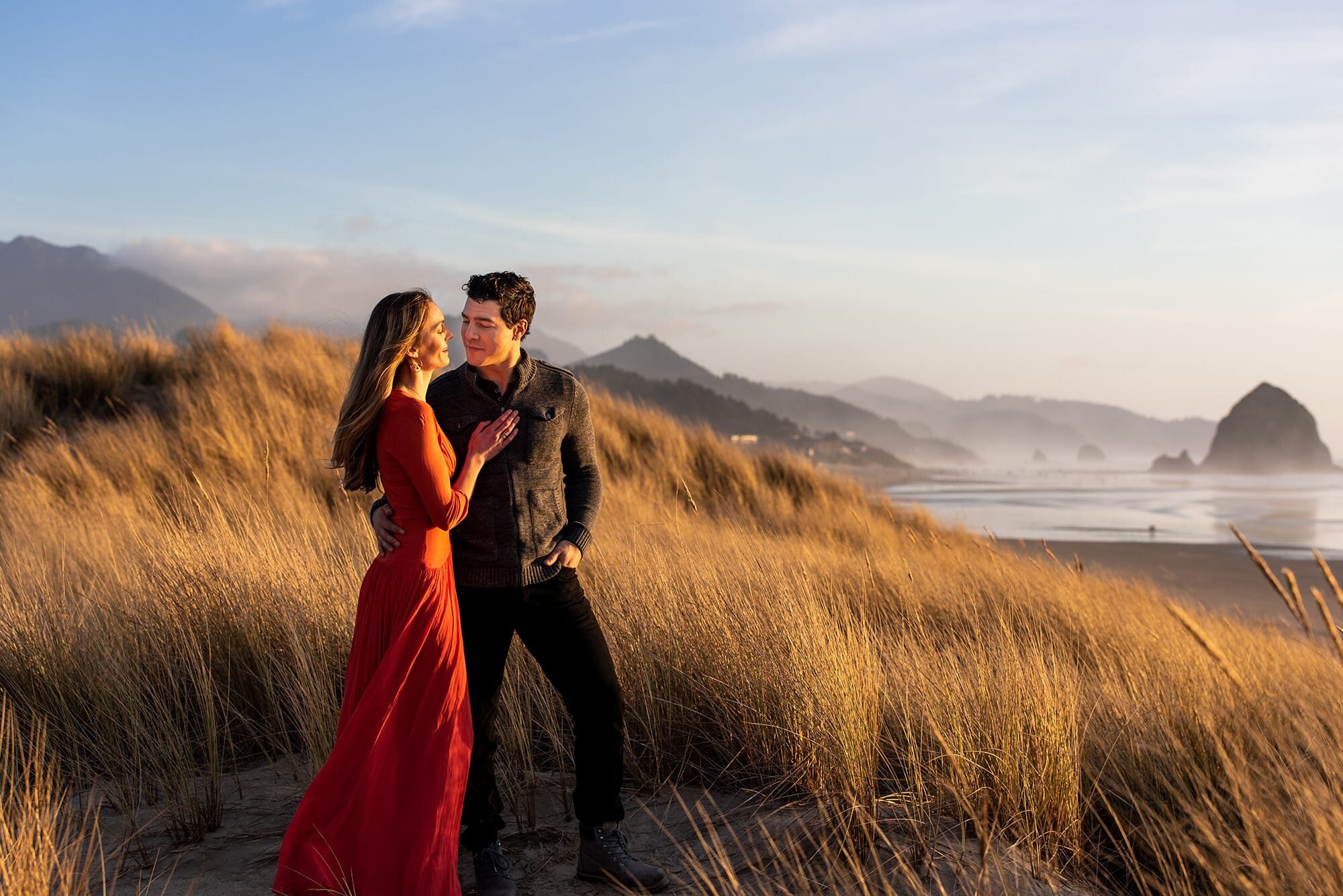 Engaged couple standing in beach grass with Haystack Rock in background