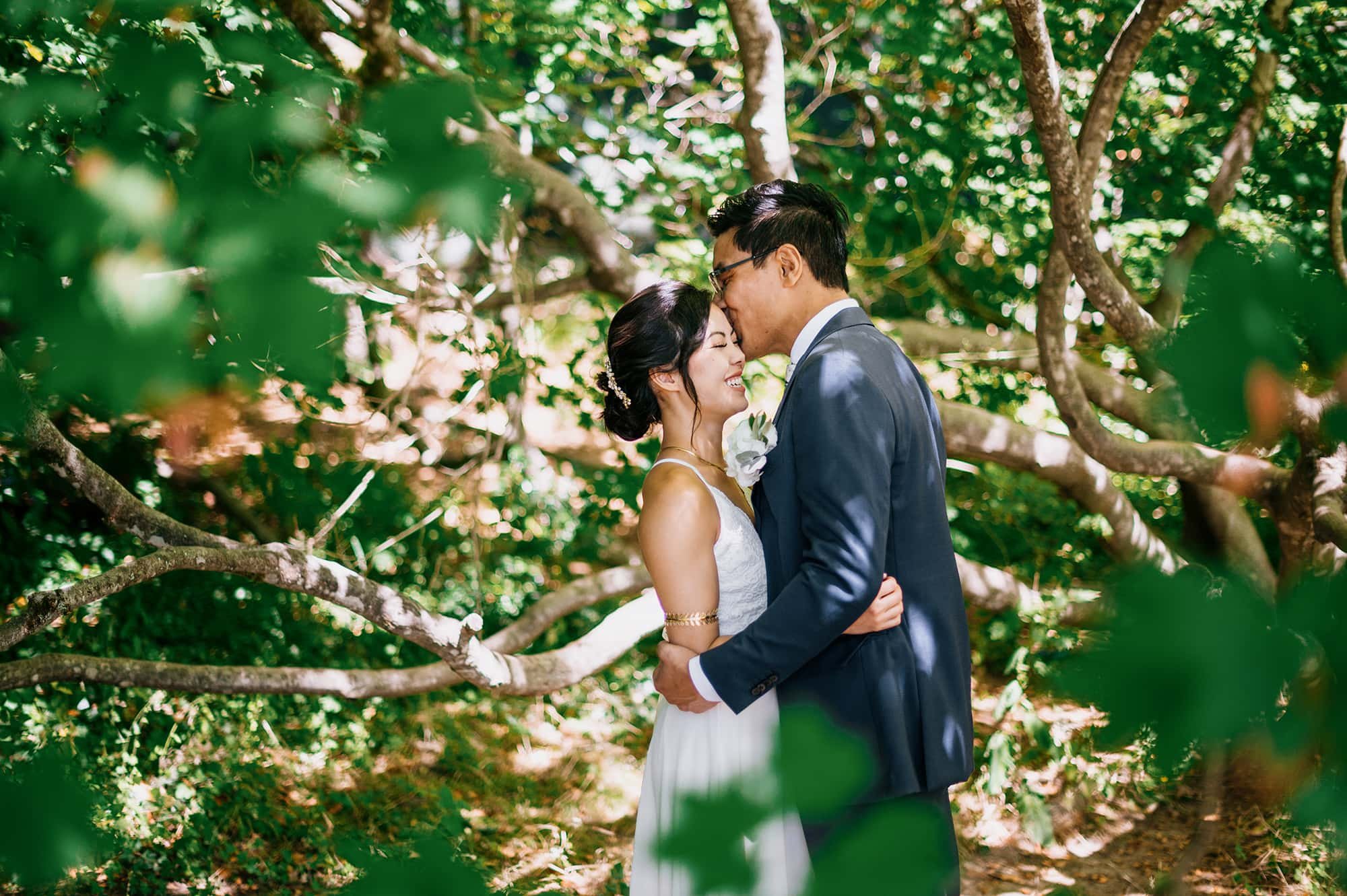 Groom kissing bride on forehead surrounded by trees