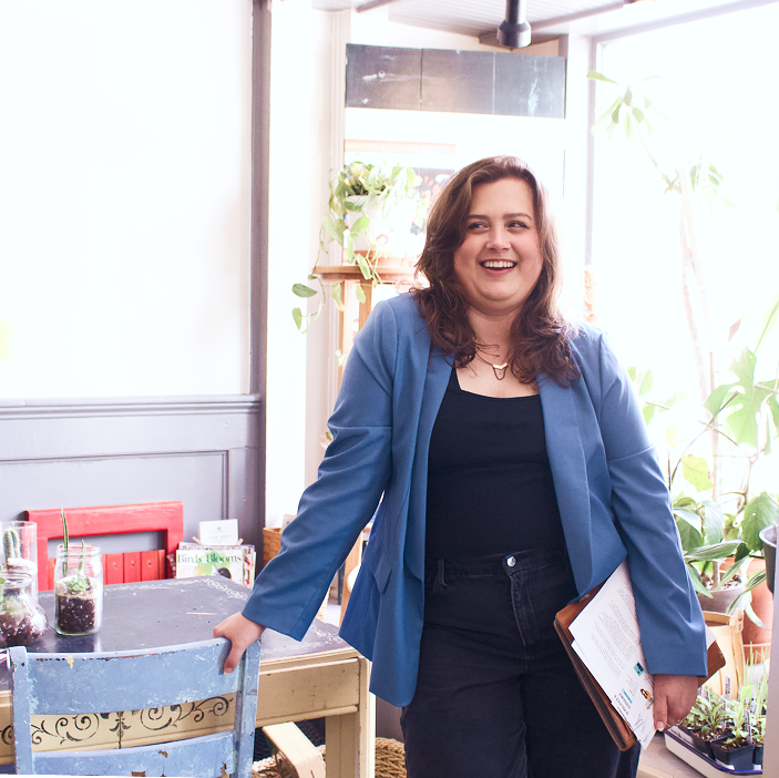 Rikki, a young white woman with curly shoulder-length brown hair, smiles while looking away from the camera, holding a Filofax and papers, wearing a blue blazer over a black tank and jeans, with plants in the background.
