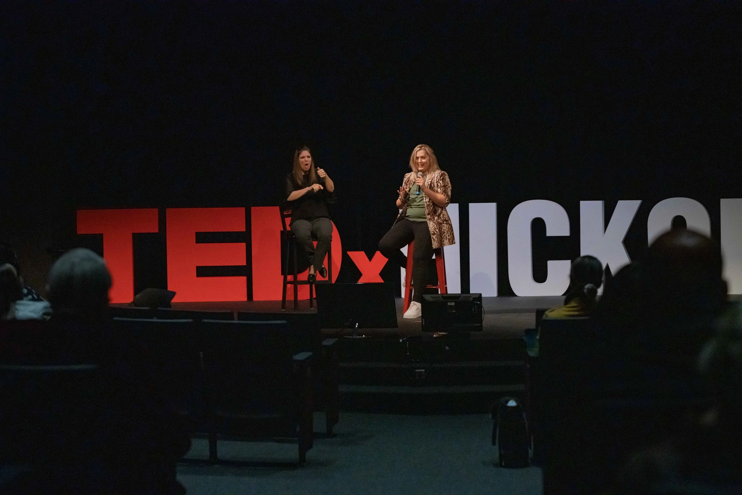 Rikki, a young white woman with shoulder-length blonde hair, speaks on a TEDx stage holding a mic, wearing a green top, black pants, and a leopard blazer, with an ASL interpreter beside her and a ‘TEDxHickory’ sign behind them.