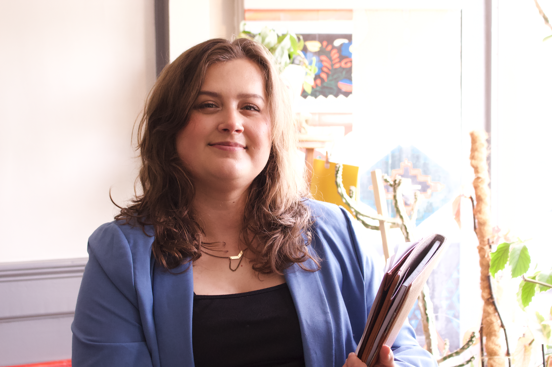 Rikki, a young white woman with shoulder-length wavy brown hair, smiles while standing in a plant shop, wearing a blue blazer over a black tank with gold jewelry and holding a Filofax.