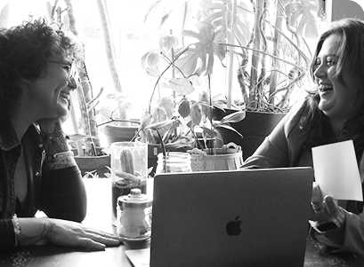 A young Latina woman with short curly hair, glasses, and wearing denim laughs with Rikki, a young white woman with her hair down. They look at each other with a laptop between them; the photo has a green overlay.