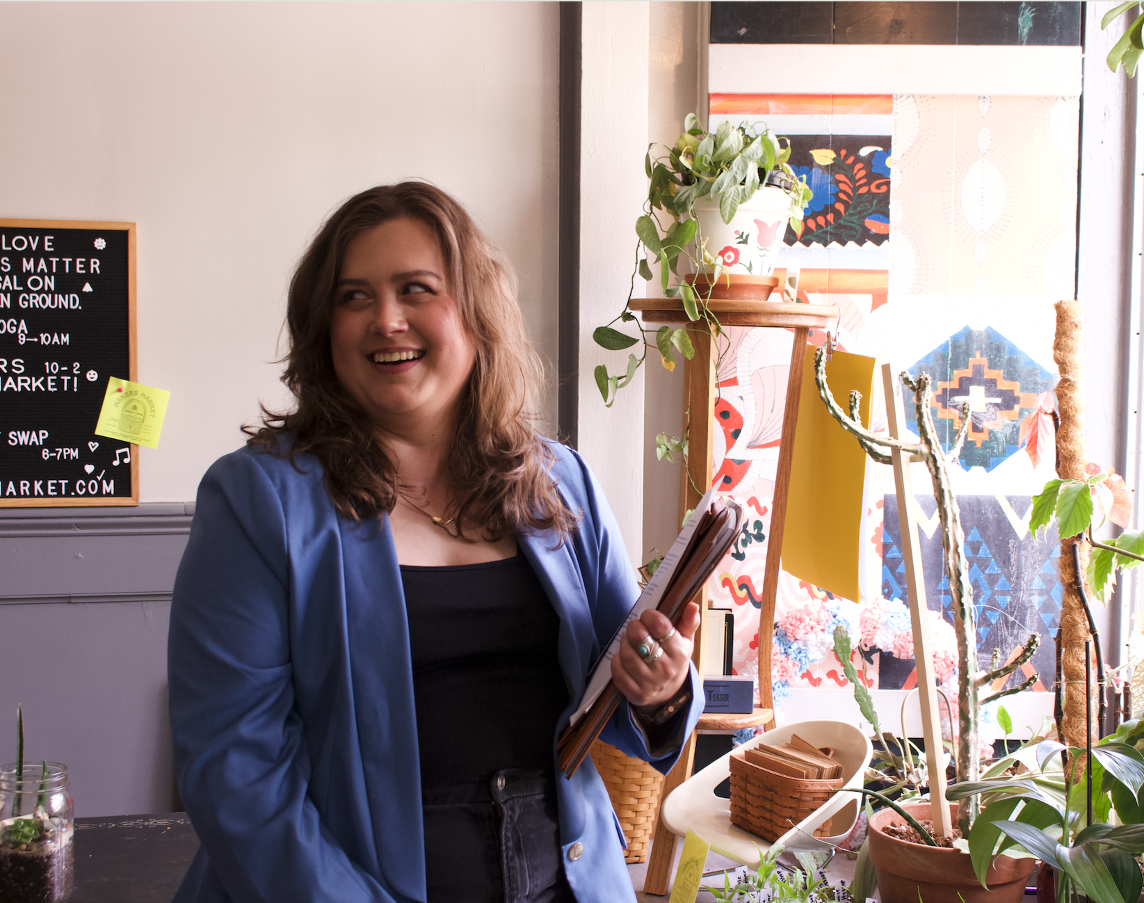 Rikki, a young white woman with curly shoulder-length brown hair, smiles while looking away from the camera, holding a Filofax and papers, wearing a blue blazer over a black tank and jeans, with plants, artwork and baskets in the background.