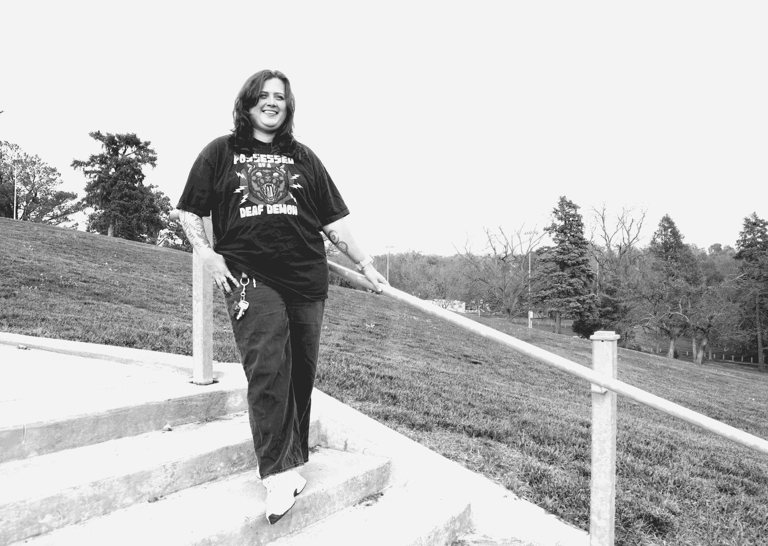 Rikki, a young white woman with shoulder-length wavy brown hair, smiles on outdoor stairs, wearing a black T-shirt with a screaming cat in hearing aids and the text ‘Possessed by a deaf demon,’ with trees and park greenery behind her.