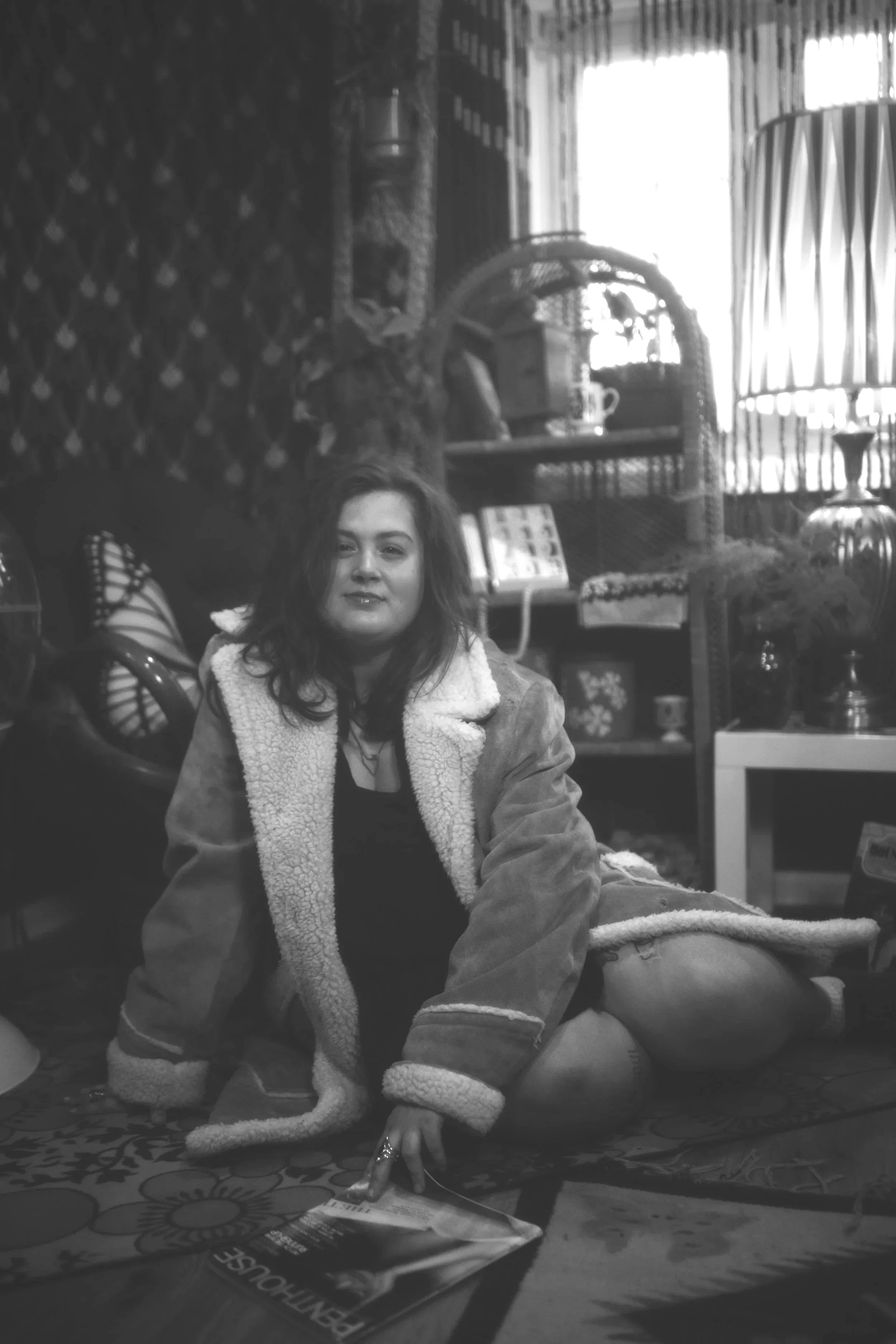 Rikki, a young white woman with wavy brown hair, wearing minimal makeup. She is wearing a black dress with a tan suede jacket. She is sitting on the floor of a vintage set. The photo is greyscale.