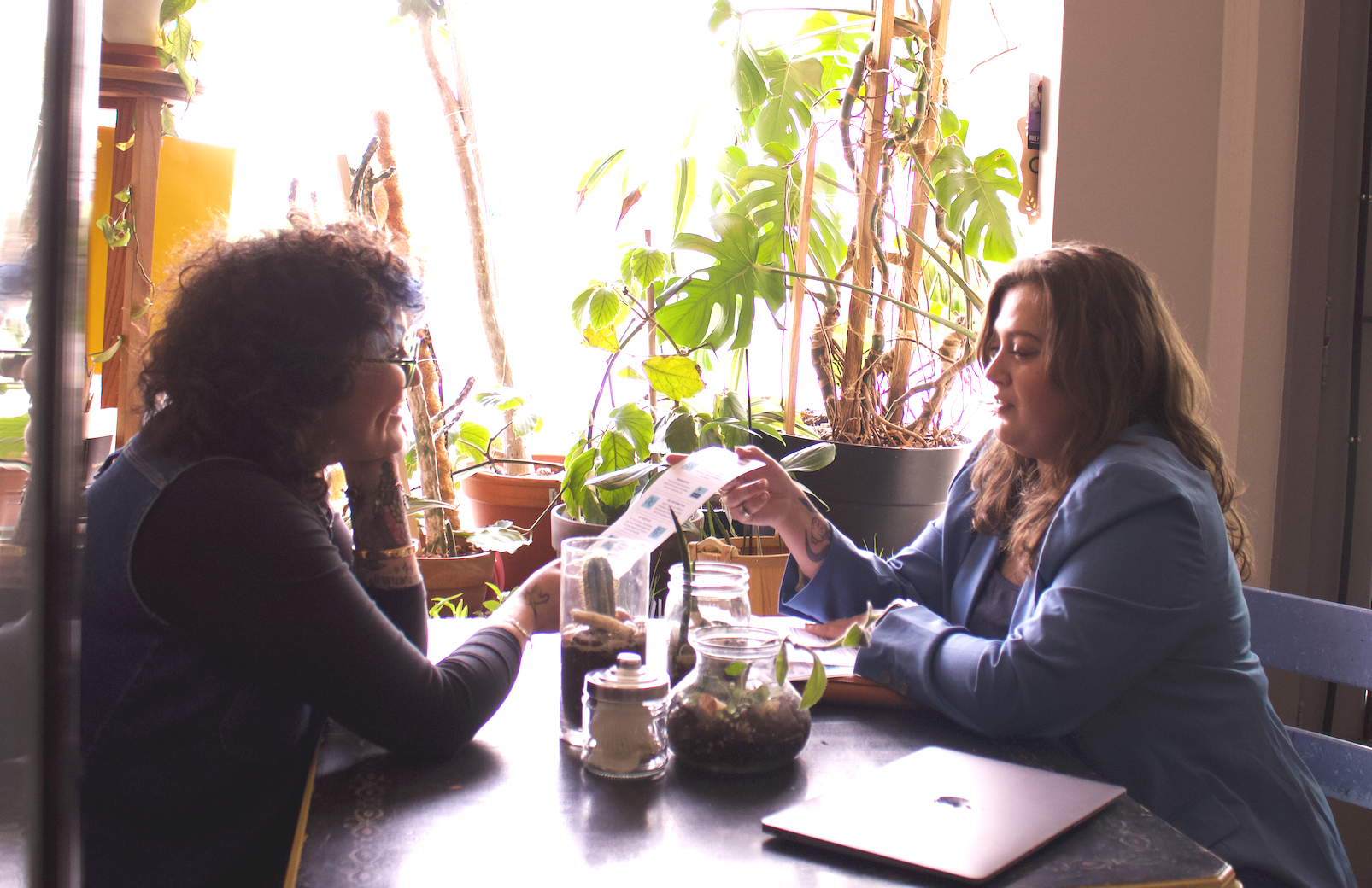 A young Latina woman with short curly hair, glasses, and wearing denim sits at a table in a plant shop with Rikki, a young white woman with her hair down. They look at each other mid-conversation as Rikki hands her an infographic.