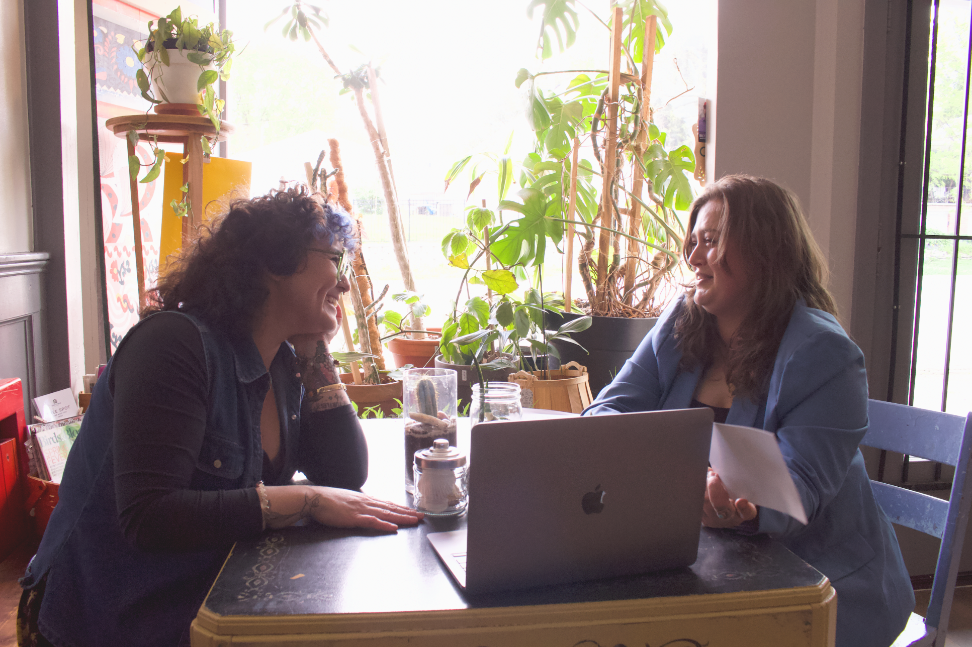 A young Latina woman with short curly hair, glasses, and wearing denim laughs with Rikki, a young white woman with her hair down. They look at each other with a laptop between them. Rikki is holding papers.