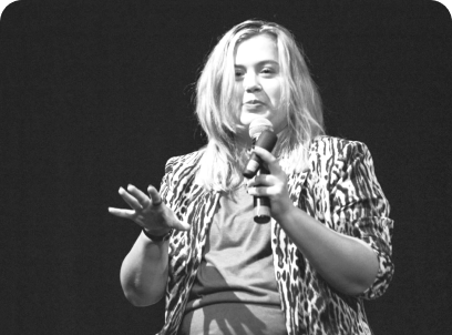 Rikki, a young white woman giving a presentation, holding a microphone and remote. She has shoulder length straight blonde hair. She is wearing a green top and leopard blazer. The photo has a green overlay.