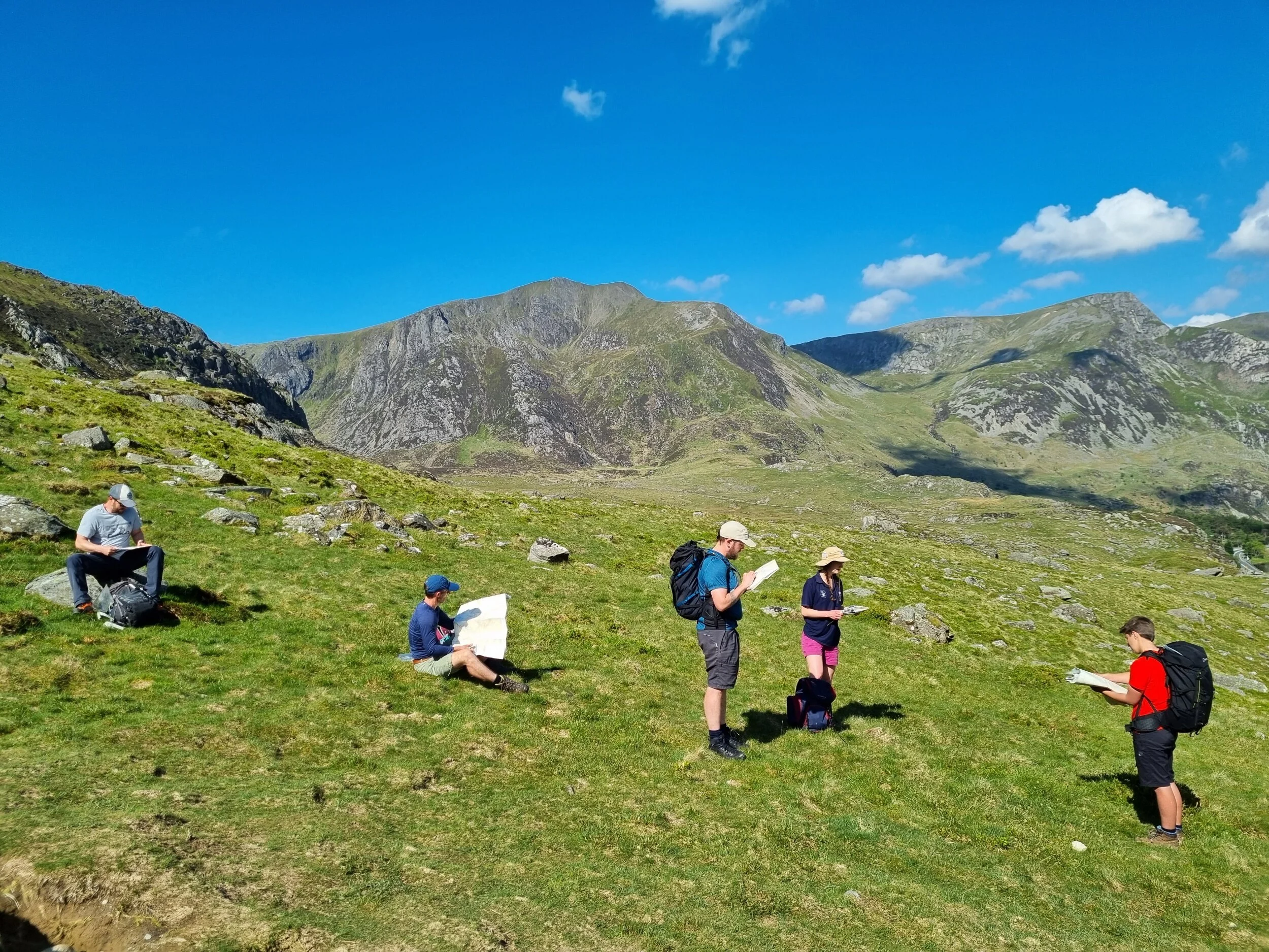 Group of six hikers reading maps and notes on a grassy hillside with mountain landscape in the background under a blue sky.