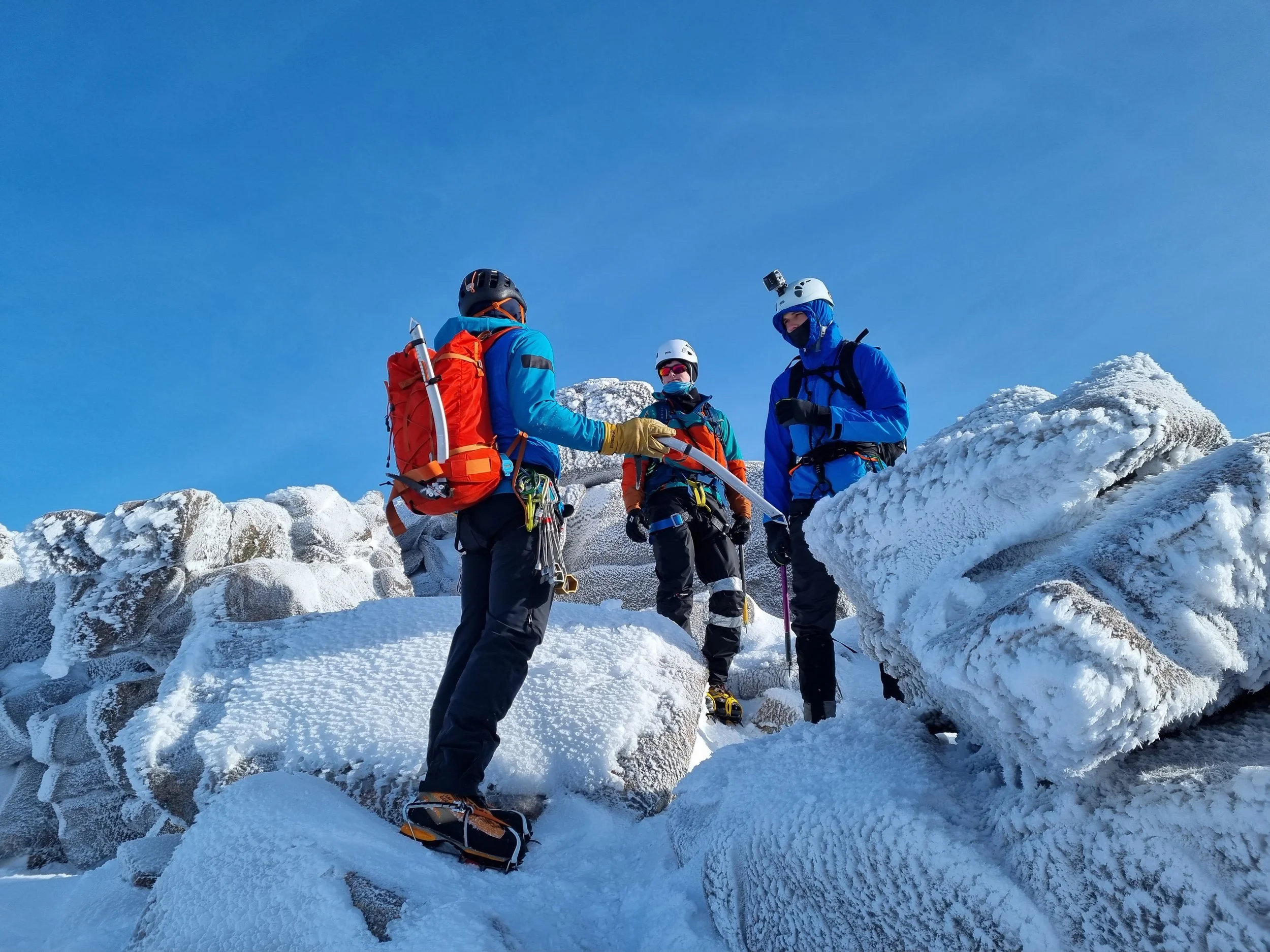 Three mountain climbers in winter gear standing on snow-covered rocks beneath a clear blue sky, with one handing an ice axe to another.