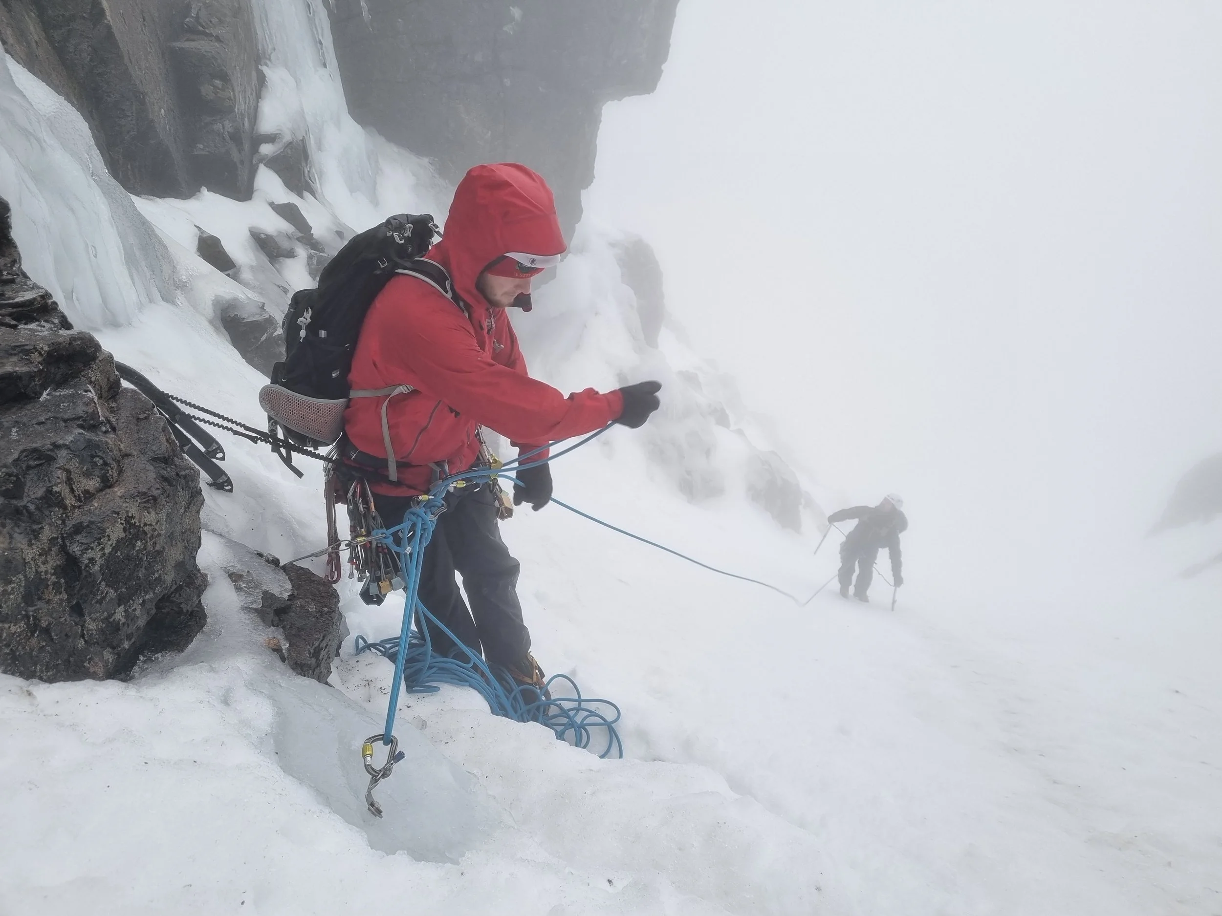 Two mountaineers climbing a snowy and icy mountain slope with ropes and climbing gear in a foggy environment.