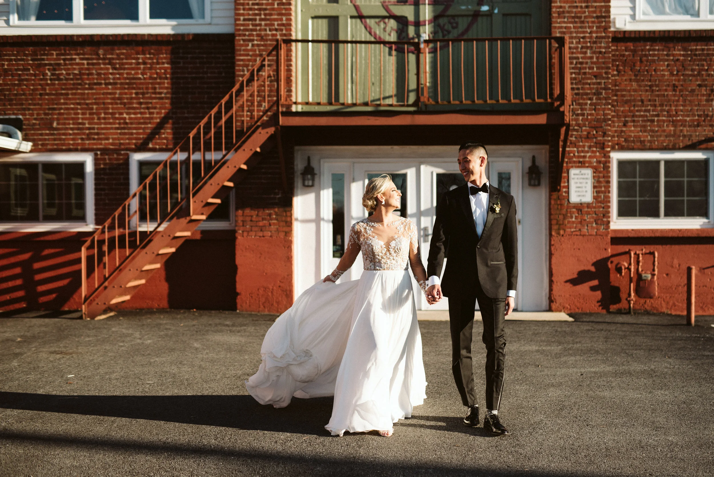 SUBJECT: The image above is a photograph of a beautiful bride with blue eyes and blonde hair, wearing a beaded, flowy white wedding dress. The groom is Chinese and is wearing a black tux. ARTIST: The photographer is Alyssa Christine Photography, who is based in Lancaster, Pennsylvania and the image was captured at the wedding location of The Reading Art Works. DETAILS: The wedding dress designer is Hayley Paige.