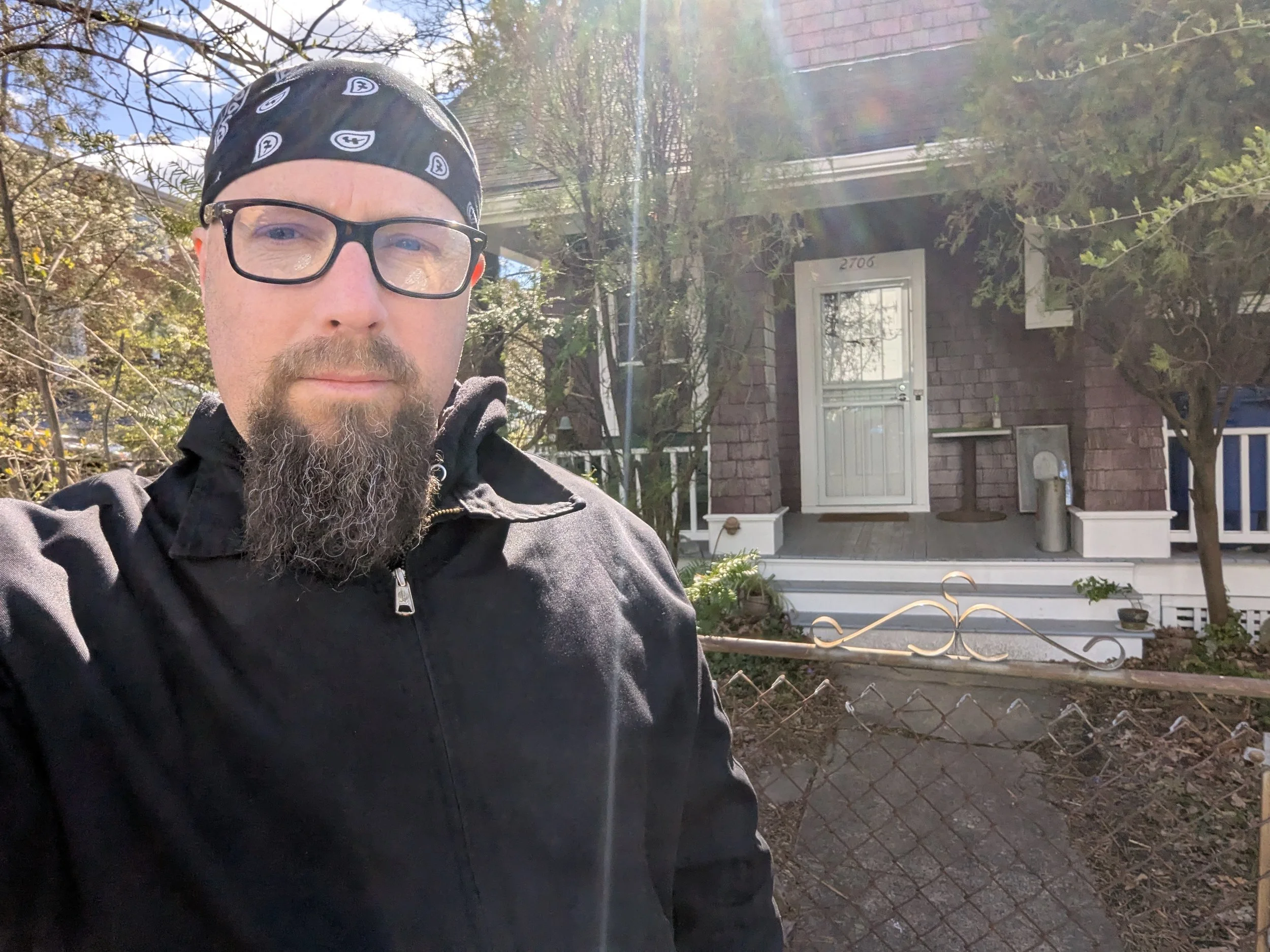 A man with glasses, a bandana, and a goatee poses in front of the Minor Threat Album cover house.