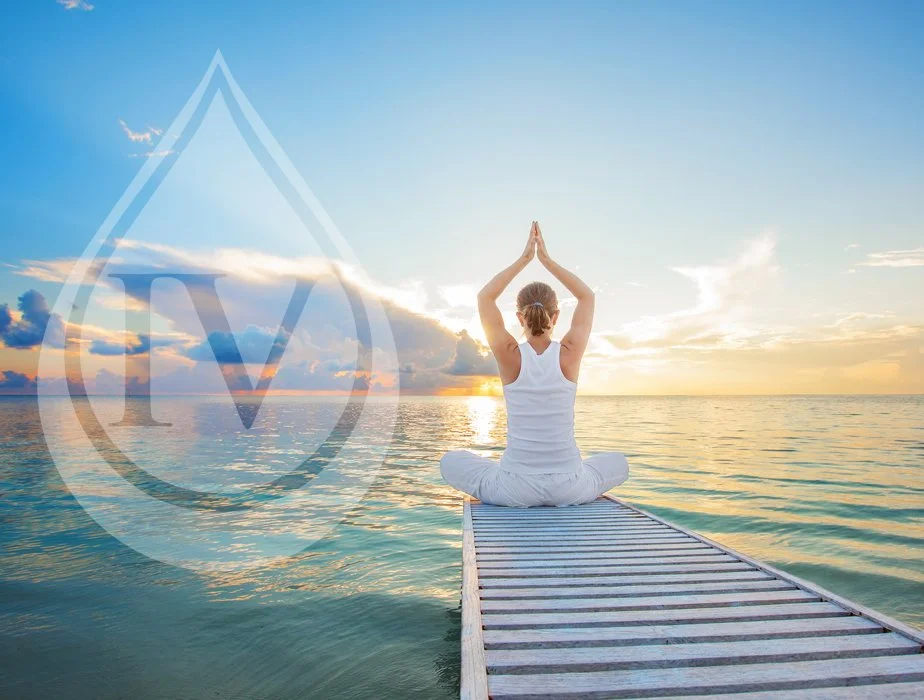 A person practicing yoga in the lotus position on a wooden pier over water during sunrise or sunset, with a calm sea and a clear sky. A transparent drop with the letter "IV" is overlaid on the image.