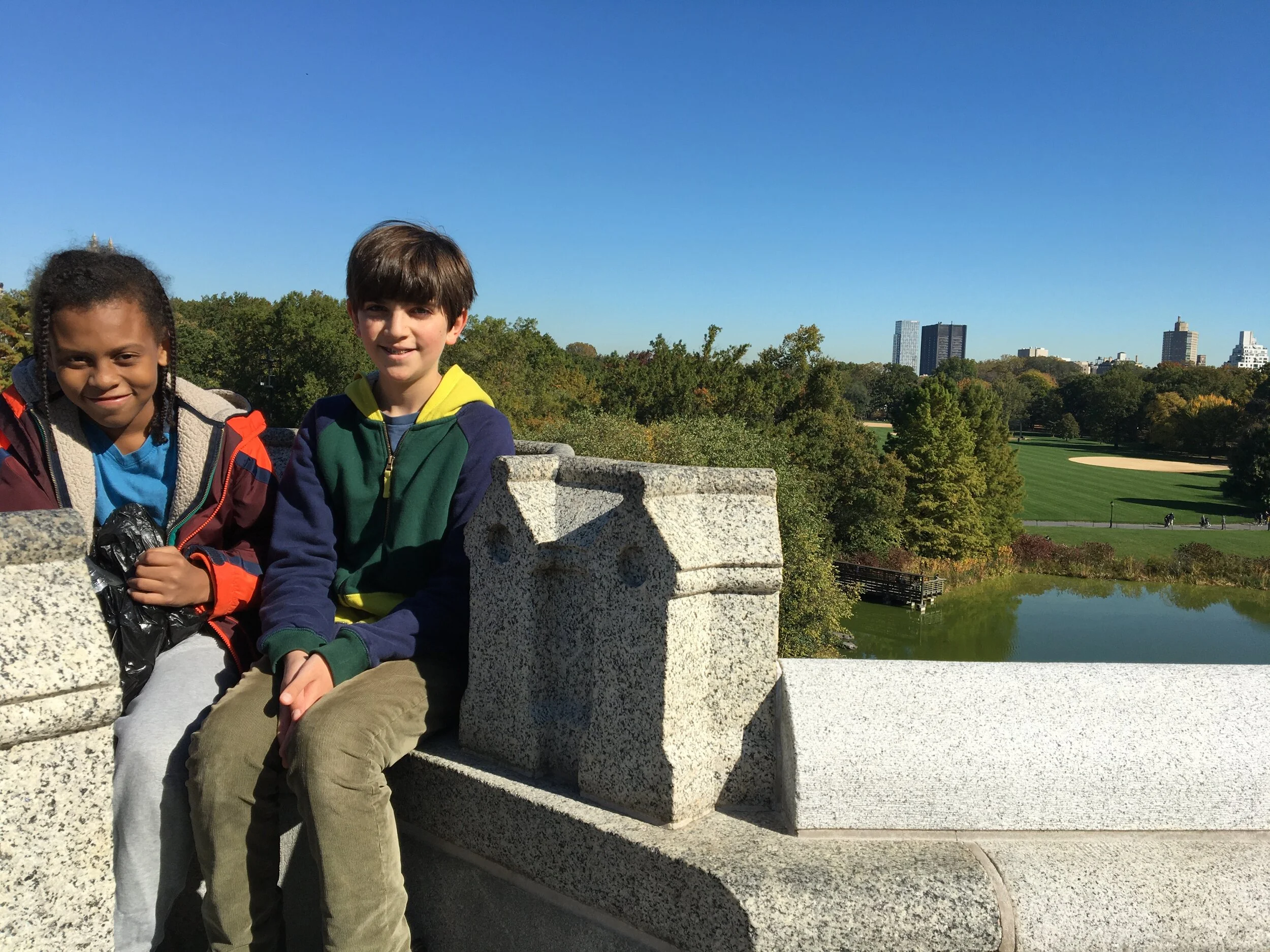 [Sterling and Hugo sitting on a stone wall, the Turtle Pond and baseball fields of central park behind them, and the city skyline in the distance.]