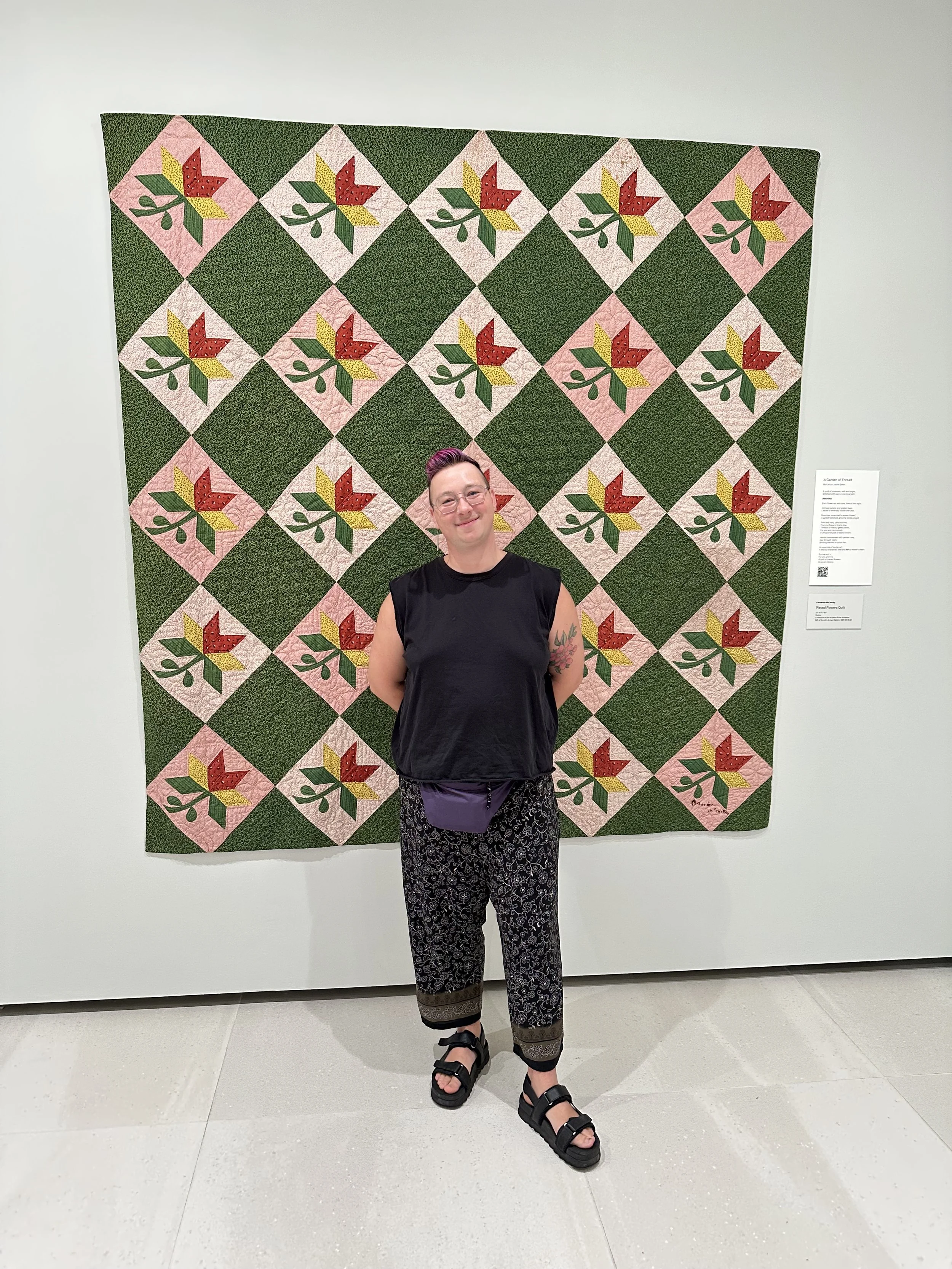 Mel, a white non-binary person with pink hair, stands in front of a quilt patterned with green and floral squares.
