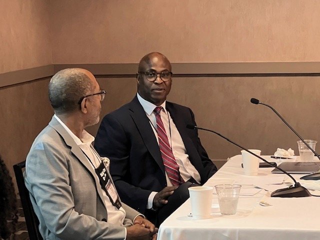 Hollis Towns and another man seated at a panel discussion table, wearing suits, having a conversation. Microphones and cups are visible on the table.