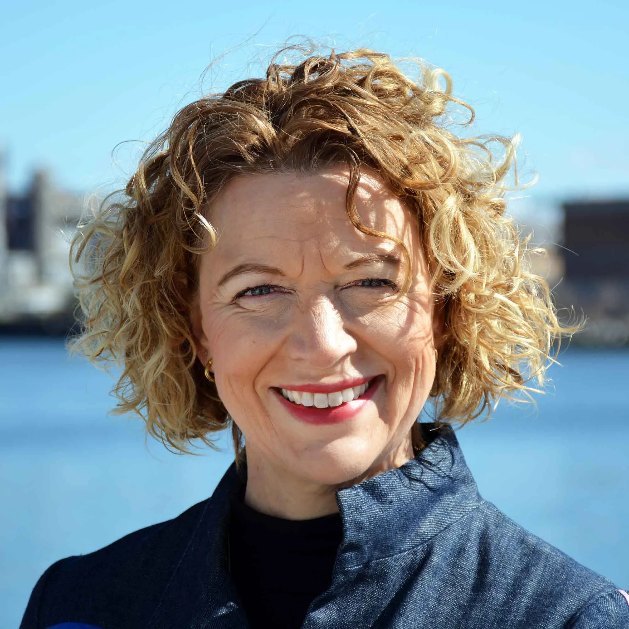 Smiling person with curly blond hair wearing a blue jacket outdoors, with a blurred waterfront background.