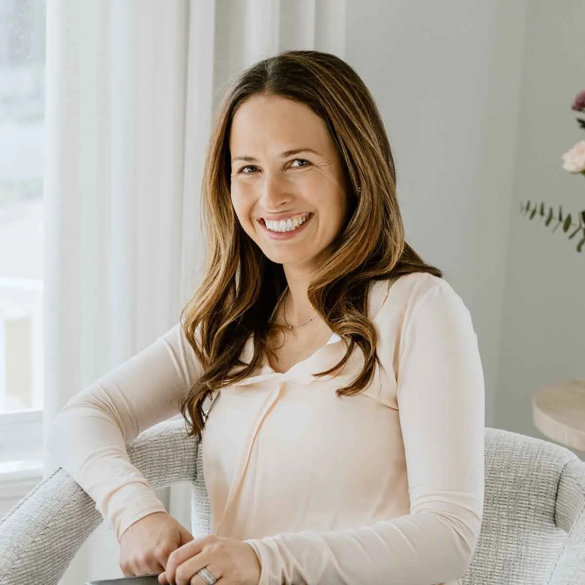 A woman sitting and smiling, wearing a light-colored long-sleeve top, with long brown hair, indoors next to a window. There are curtains and a small table with flowers in the background.