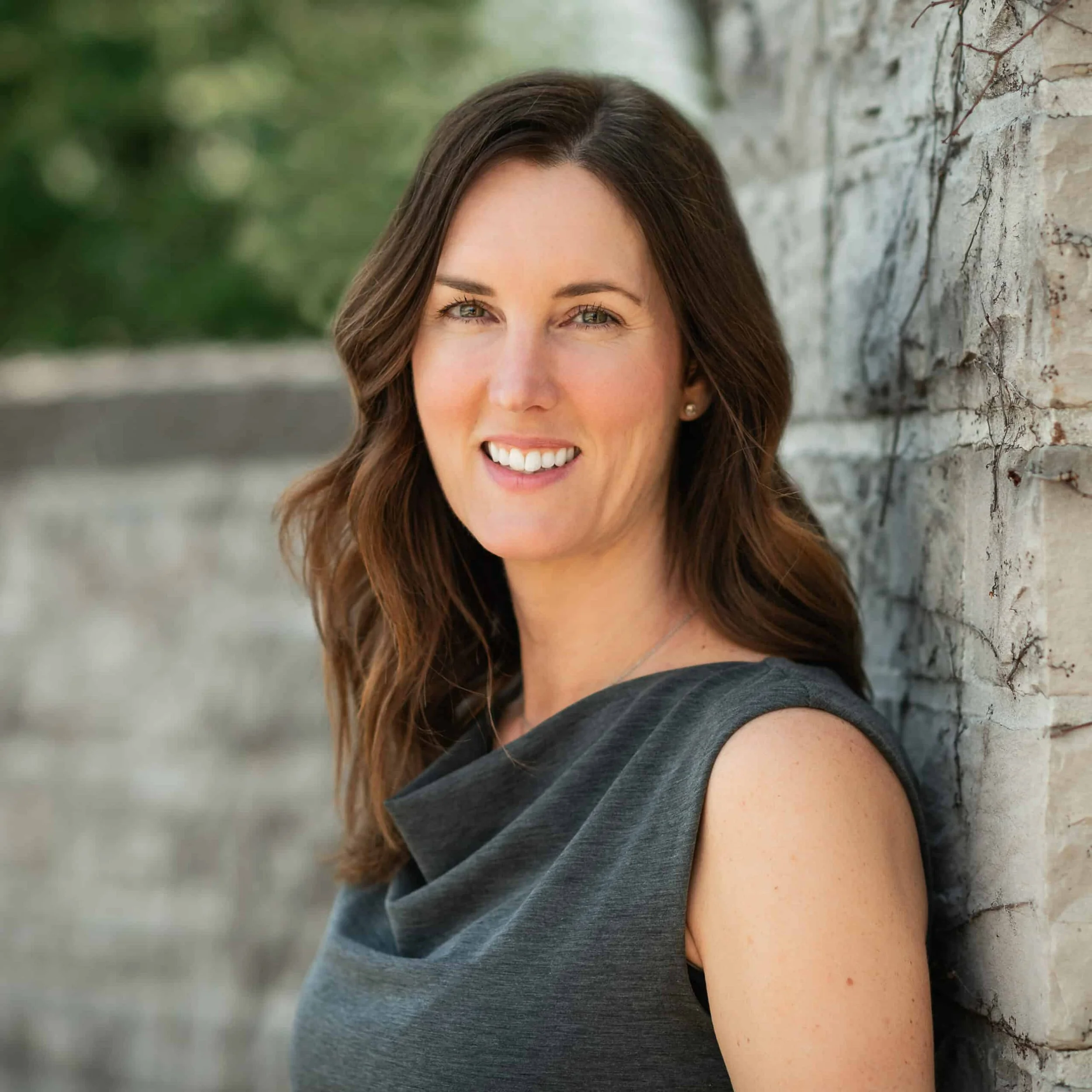 Smiling woman with long brown hair wearing a sleeveless gray dress, standing against a stone wall outdoors.