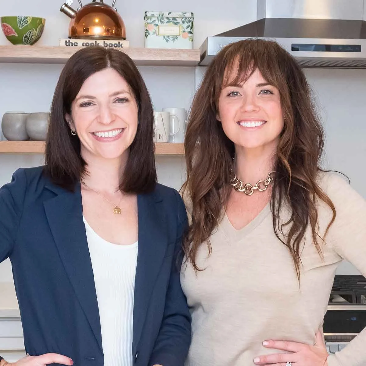 Two women smiling in a kitchen environment, with shelves holding mugs and kitchen items in the background.