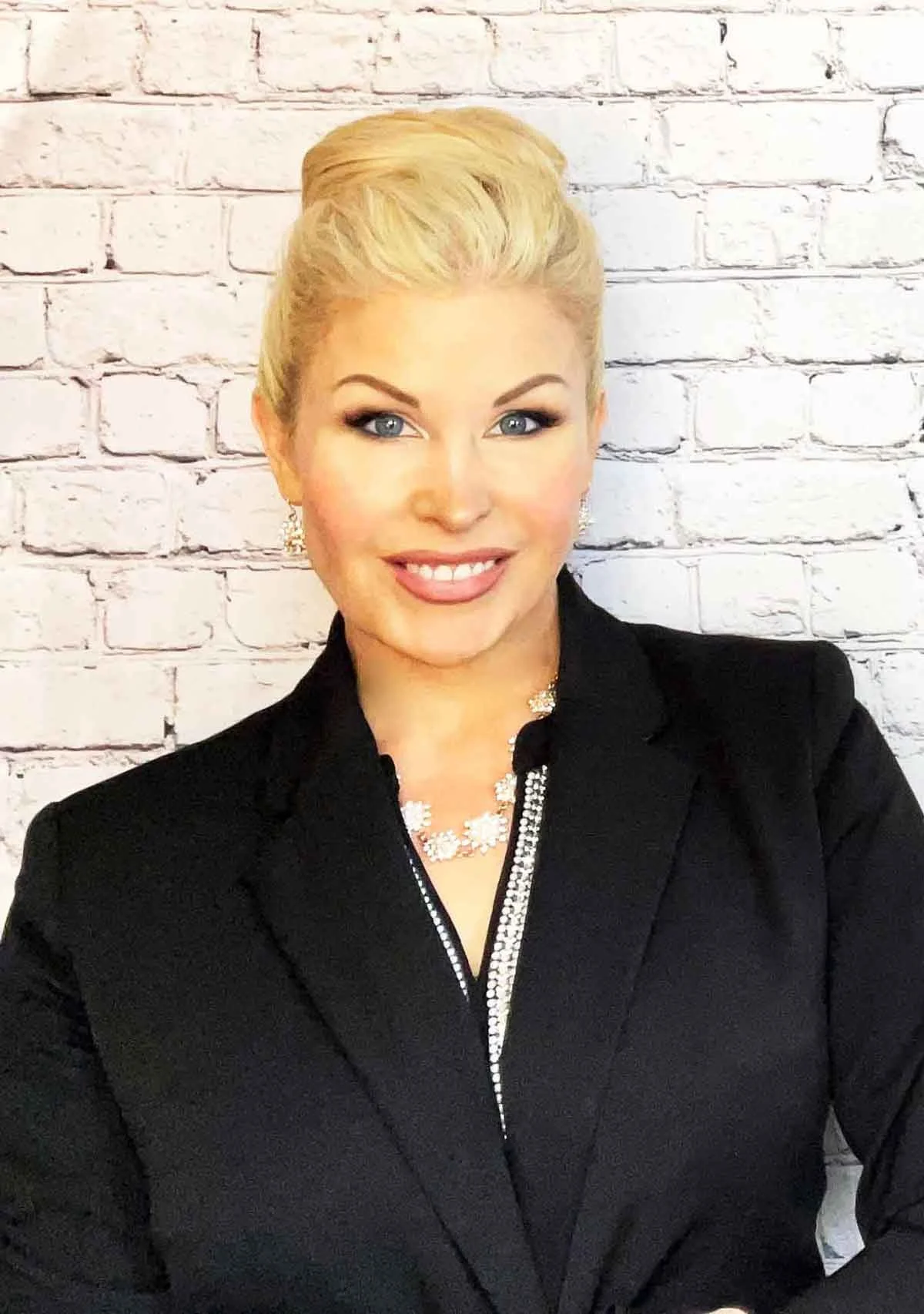 Person with blond hair and black blazer smiling in front of white brick wall