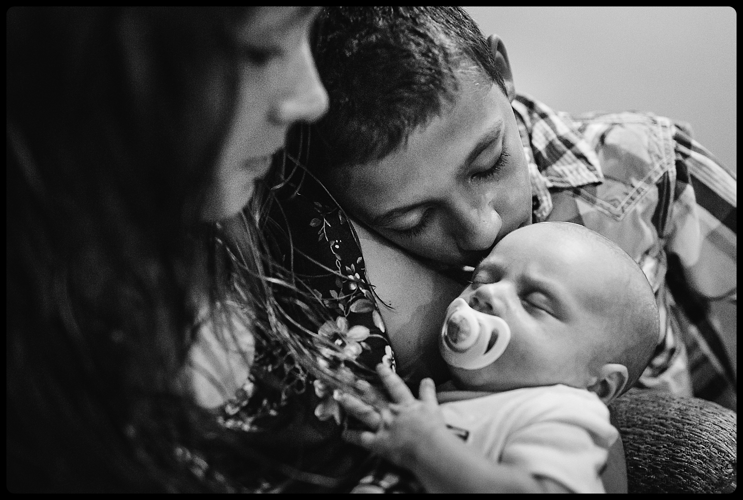 Young boy kisses newborn sibling while mother snuggles both.