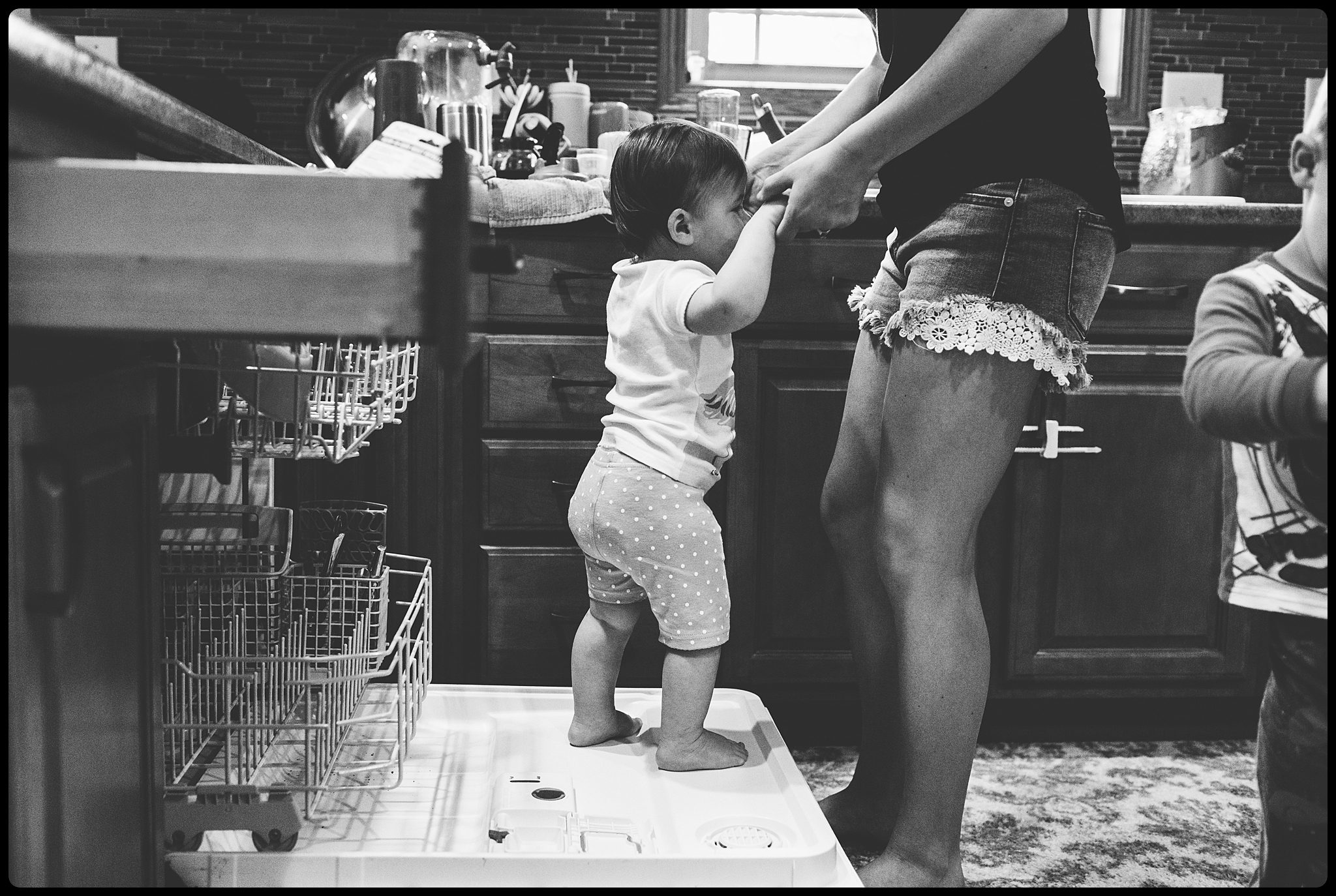 Baby climbs on dishwasher while mom is trying to clean.