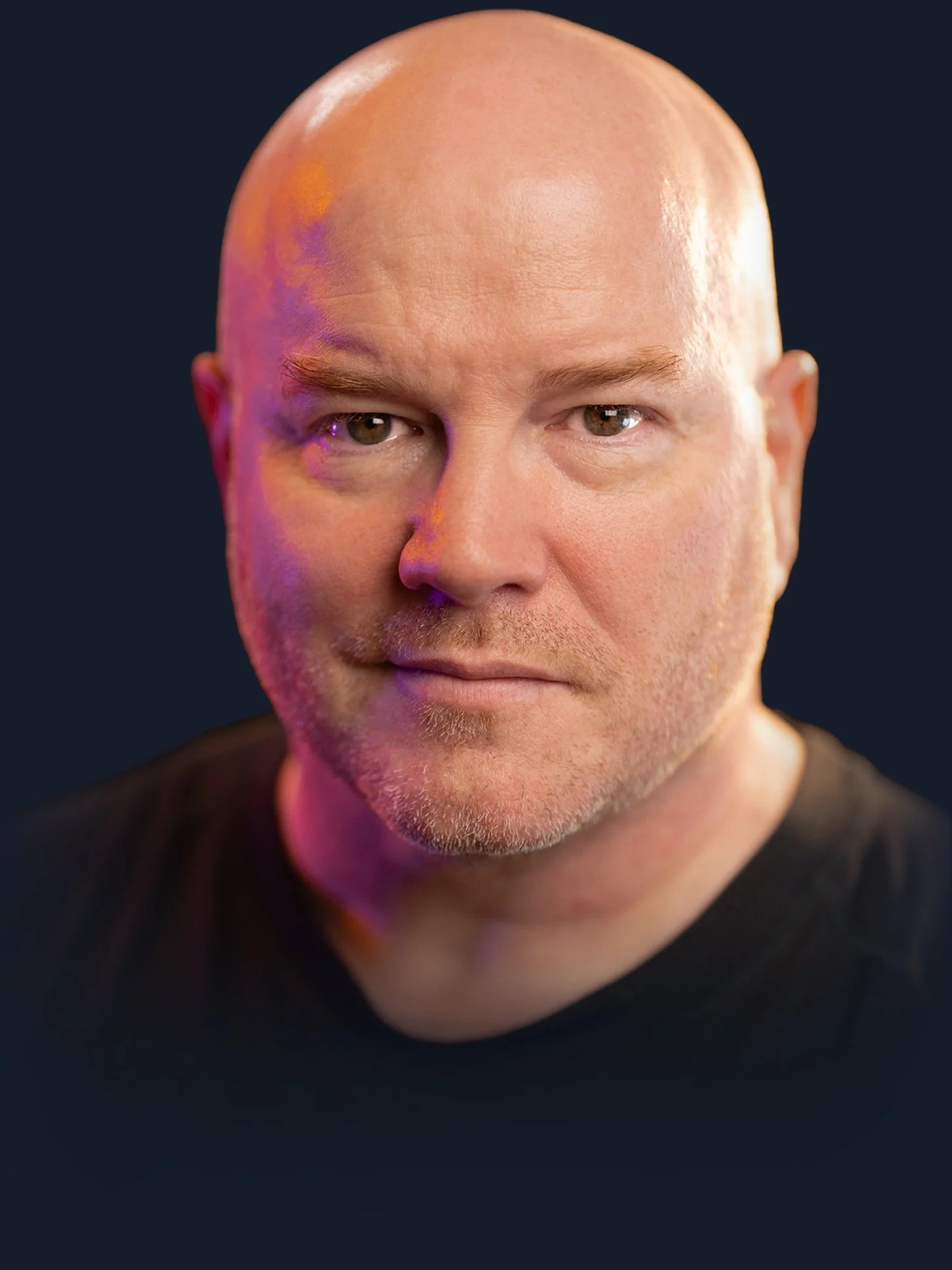 Close-up portrait of a bald middle-aged man with light skin, smiling slightly, wearing a black shirt, against a dark background with purple and orange lighting.