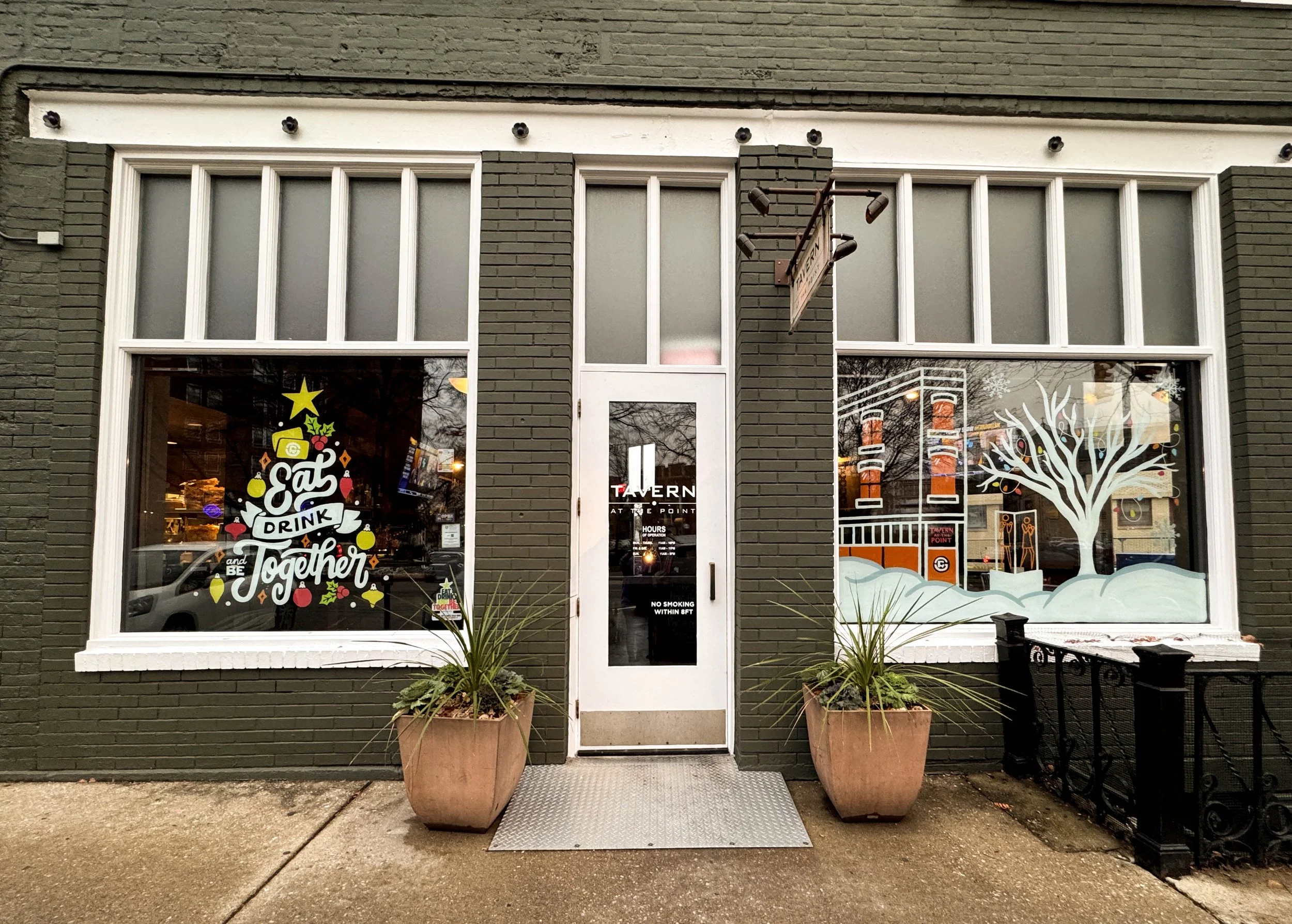 storefront with a black brick facade, decorated windows, potted plants, and an entrance door, featuring holiday-themed window art and a sign that reads 'Tavern At The Point'.