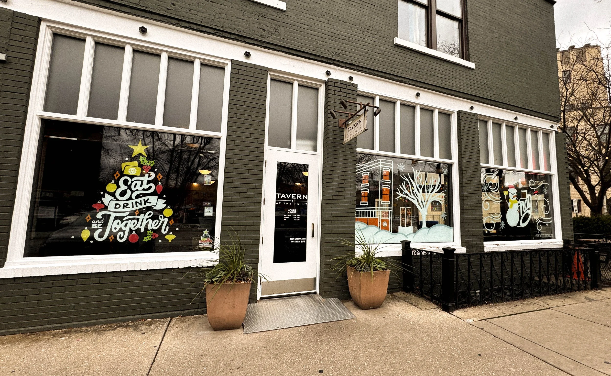 Exterior view of a restaurant with large windows decorated with holiday-themed artwork, including a Christmas tree with a star on top, a snowman, and a winter scene with snow-covered trees. The entrance has a glass door with the restaurant's name and