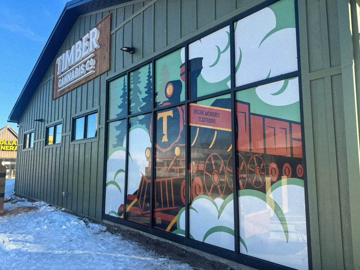 Green building with large window mural of a red steam train labeled 'High Minded Express' and a wood sign that reads 'Timber Cannabis'.