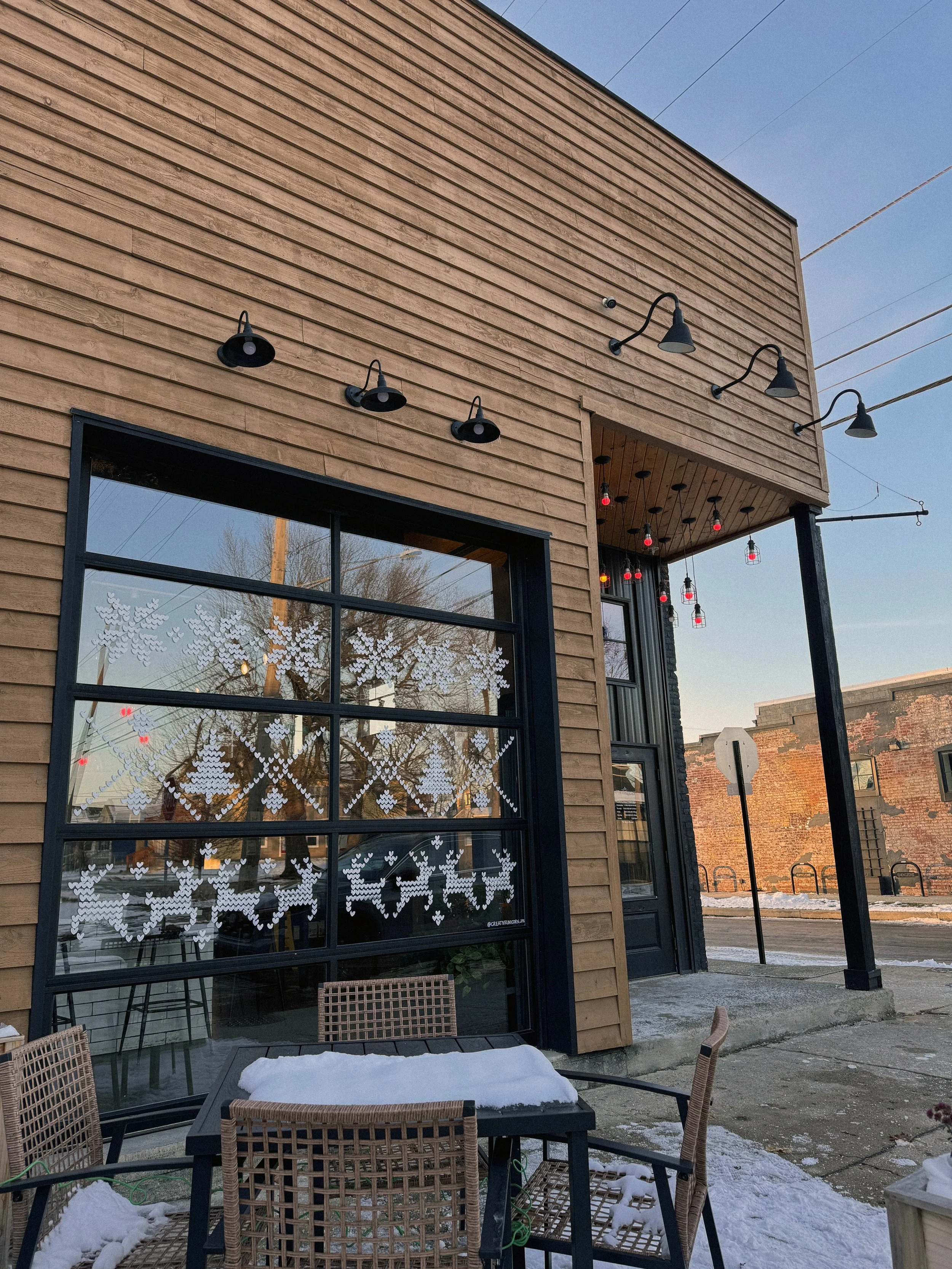 Exterior of a restaurant or cafe with holiday window decorations, outdoor seating area, and string lights against a clear sky.