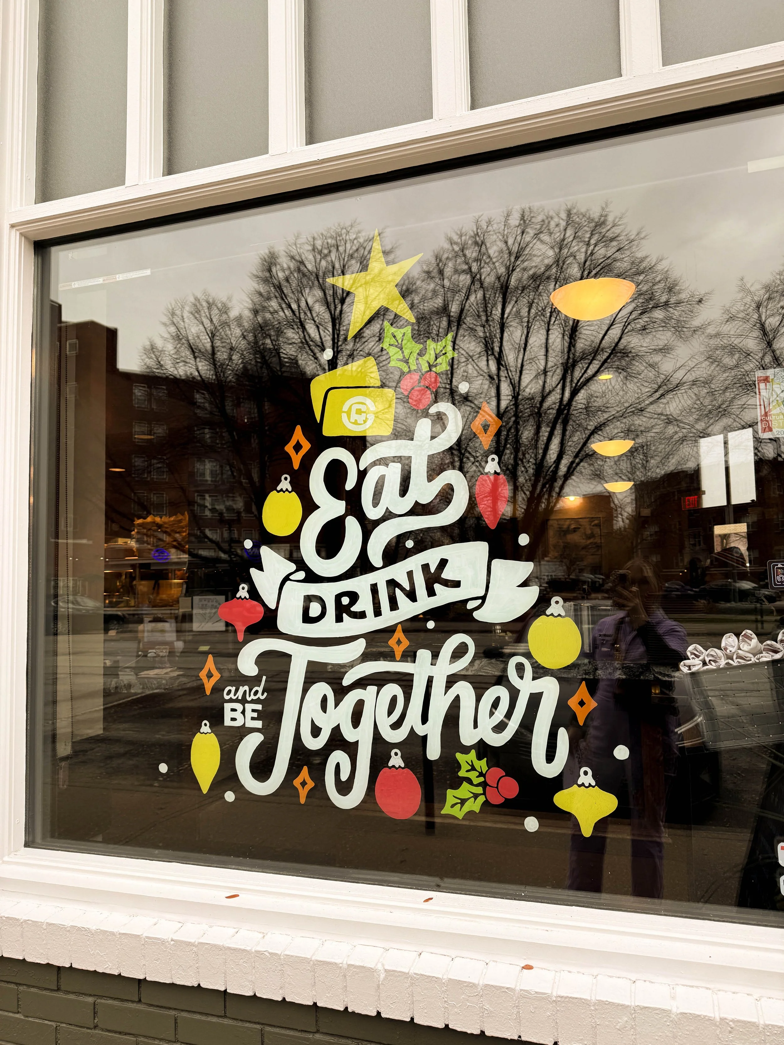Holiday-themed window display with the message 'Eat, Drink, and Be Together' decorated with Christmas ornaments, holly, berries, and stars.