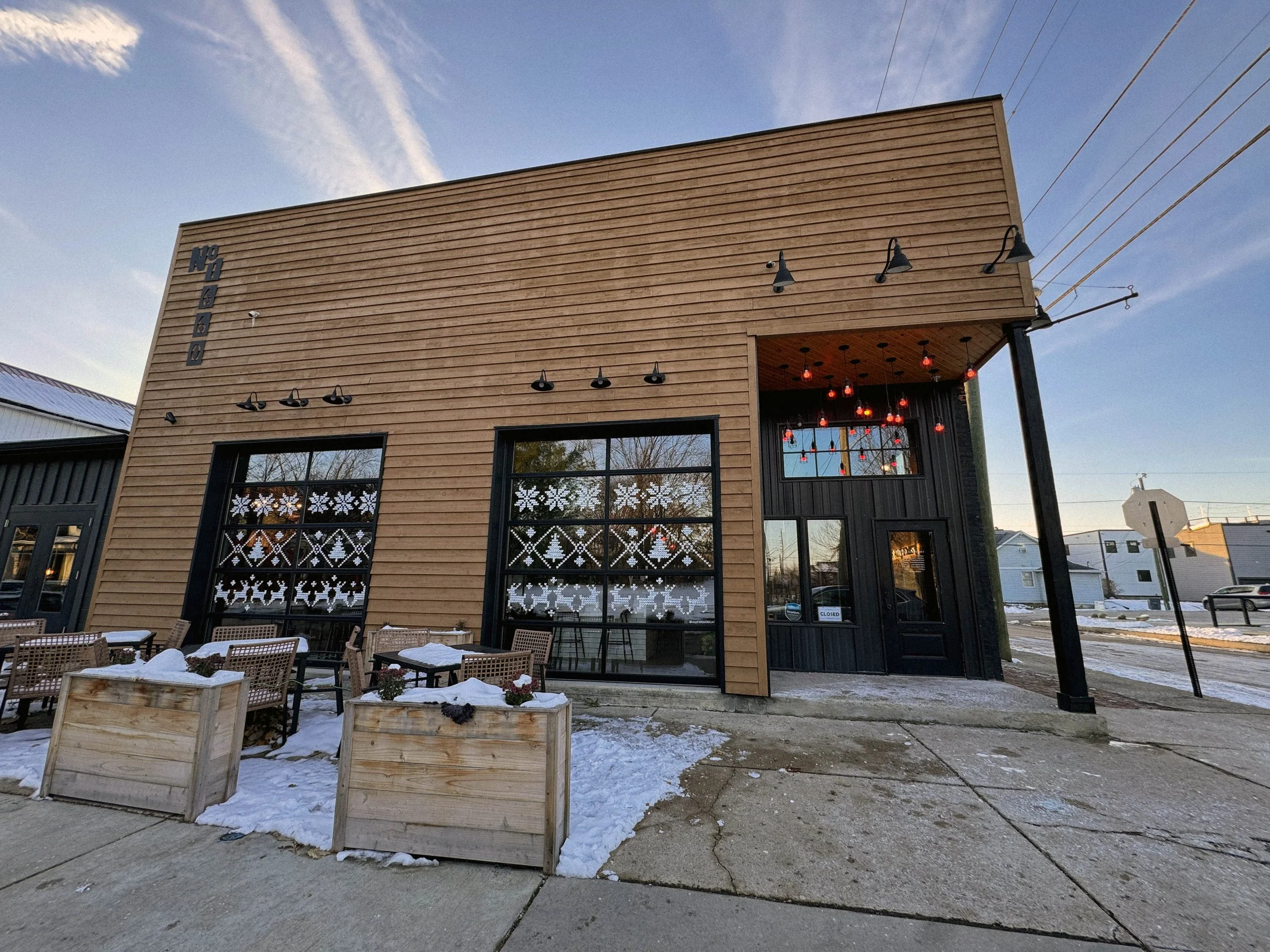 A modern storefront building with large windows decorated with snowflake patterns, displaying hanging red lights inside. The exterior features light brown horizontal siding and a black entrance door. There are wooden planters with snow and outdoor se