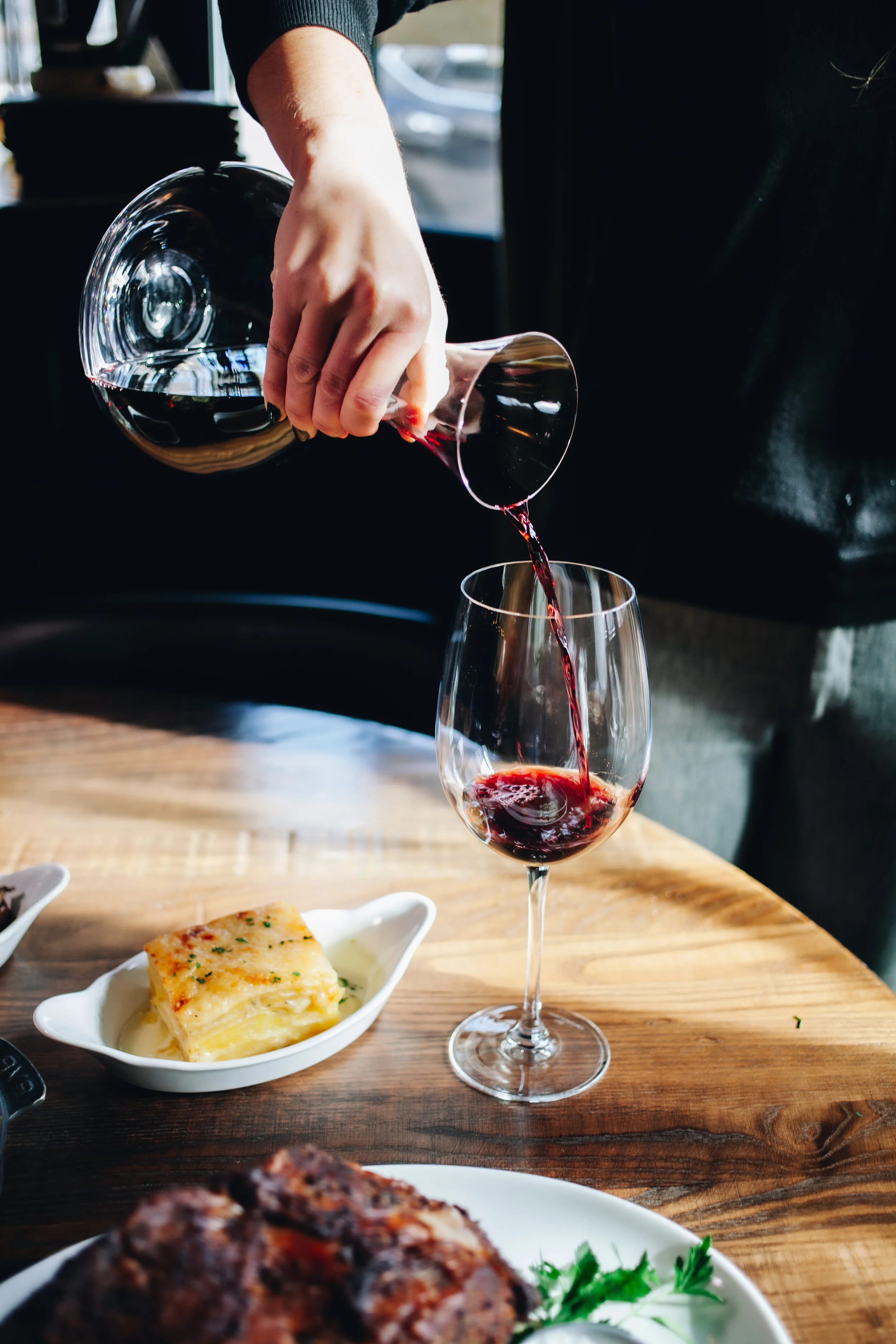 Person pouring red wine from a glass bottle into a wine glass on a wooden table. In the background, there is a plate with a baked dish and part of a dish with a grilled meat item.