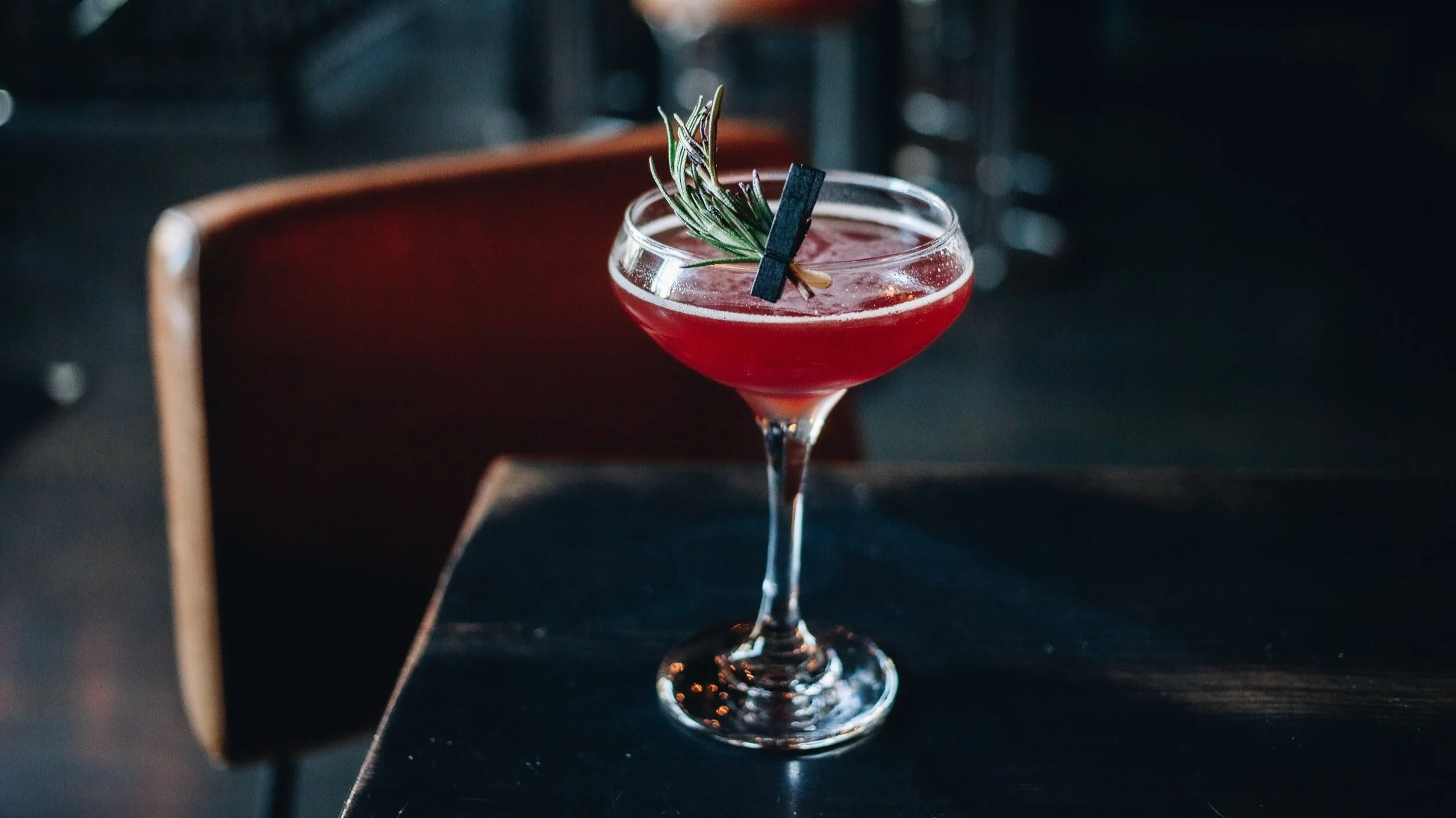 A red cocktail in a coupe glass garnished with a sprig of rosemary and a small clothespin, on a dark surface with a blurred background.