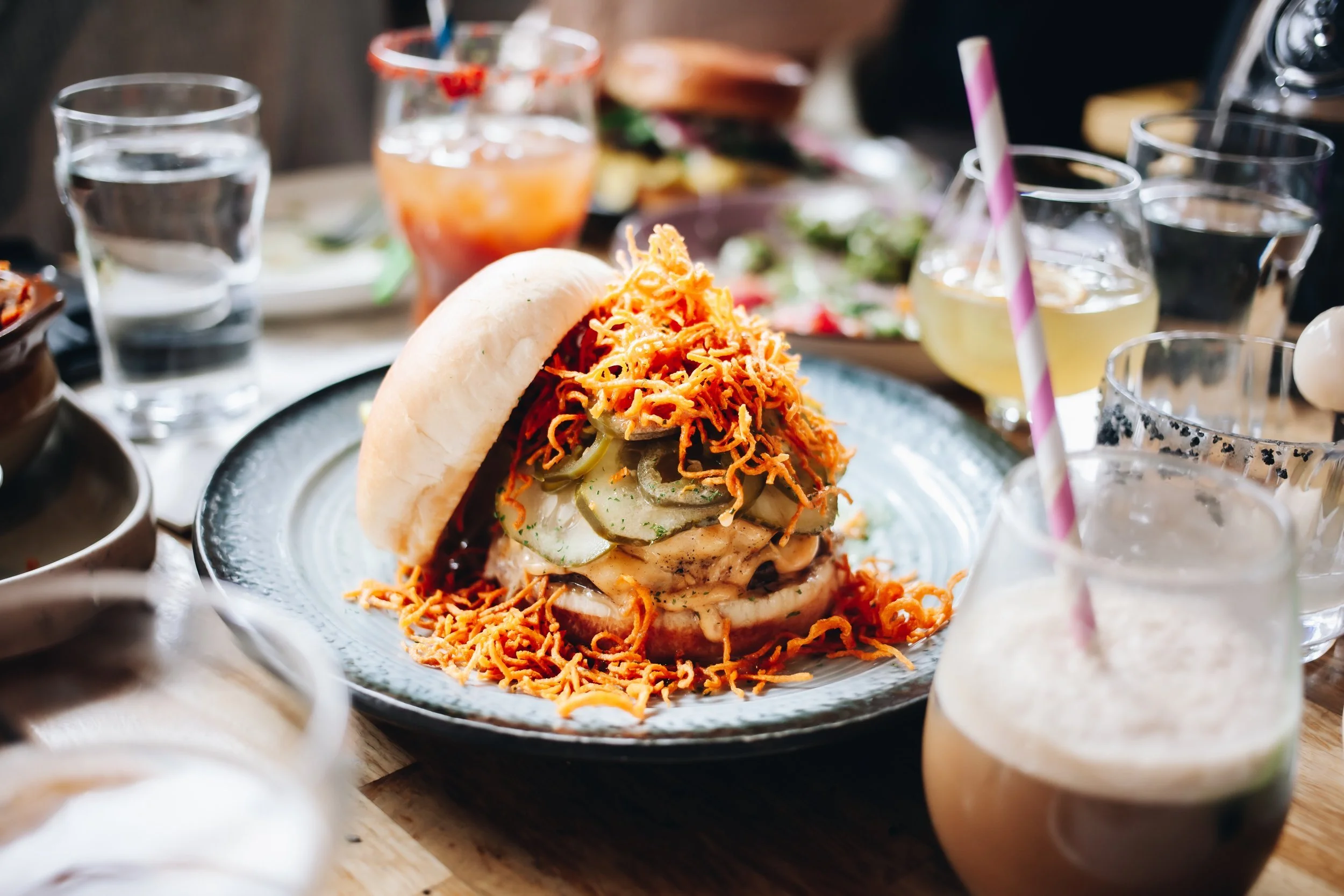A sandwich with shredded cheese, sliced pickles, and other ingredients on a bun, surrounded by glasses of iced beverages and other food items on a wooden table.