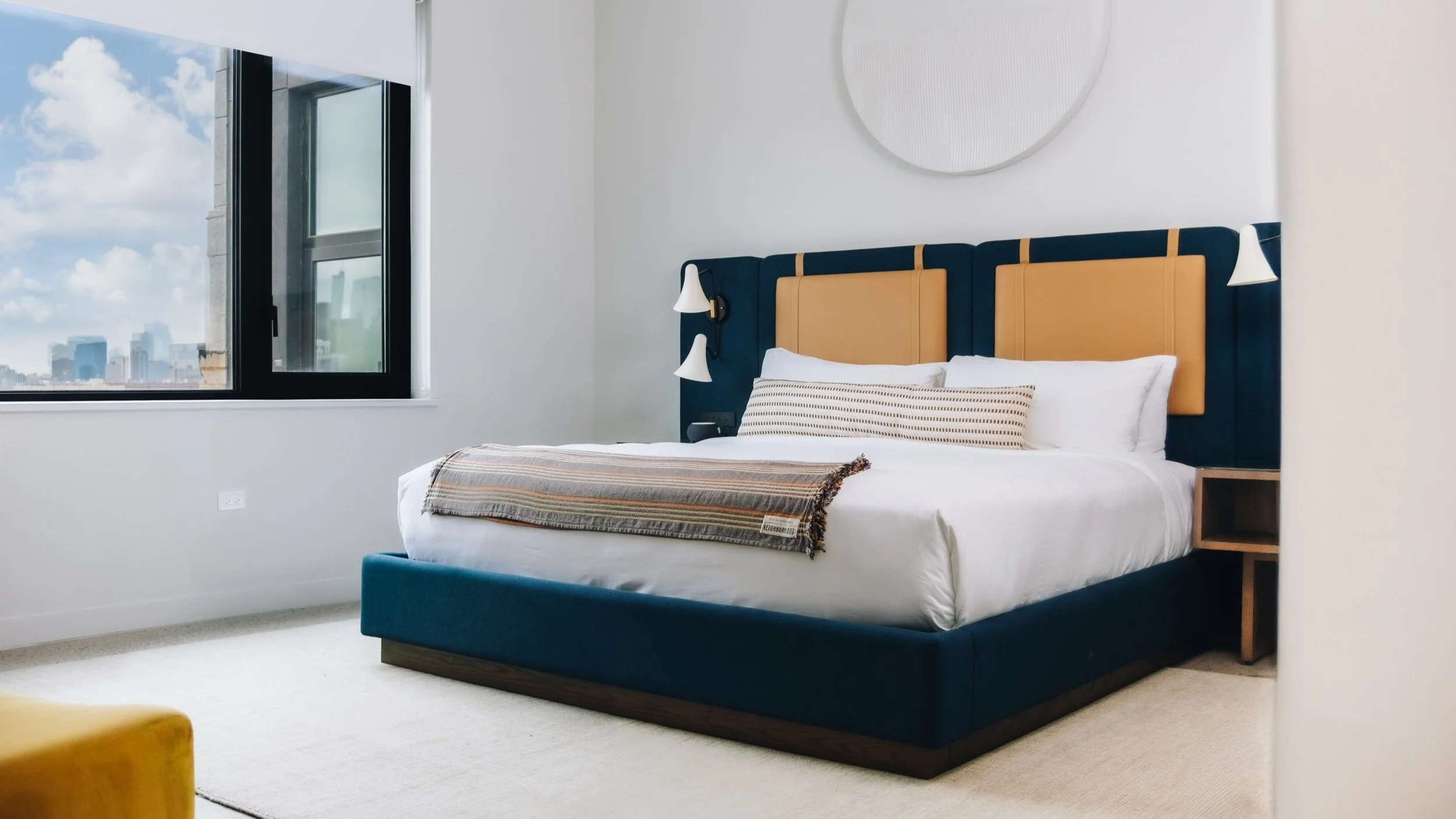 A modern bedroom with a large bed featuring a blue and orange headboard, white bedding, and pillows. Next to the bed are built-in reading lights and a nightstand. There is a large window showing a cityscape and blue sky with clouds.