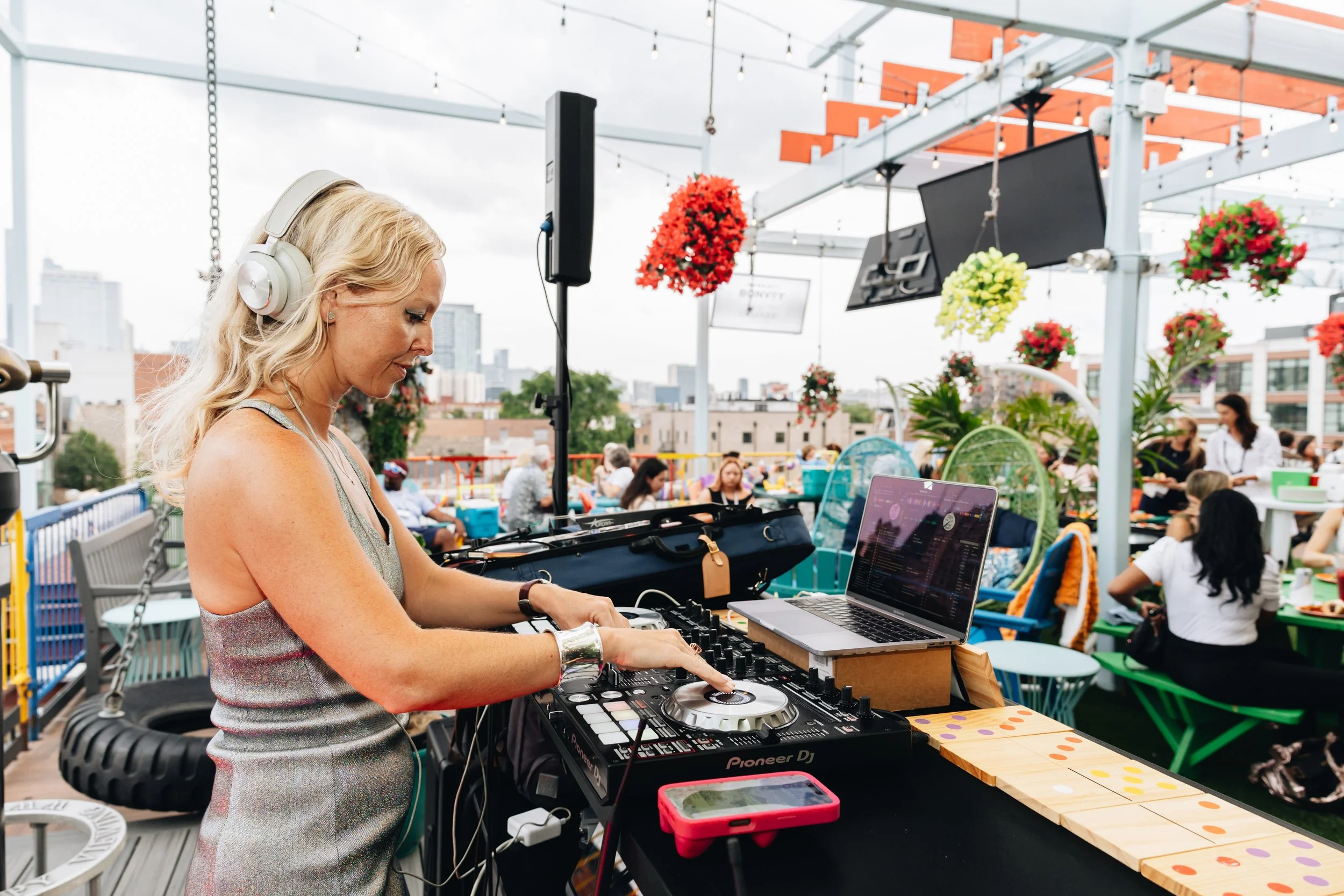 A blonde woman wearing headphones and a sleeveless dress DJing with a Pioneer DJ controller on an outdoor rooftop terrace during a daytime event.