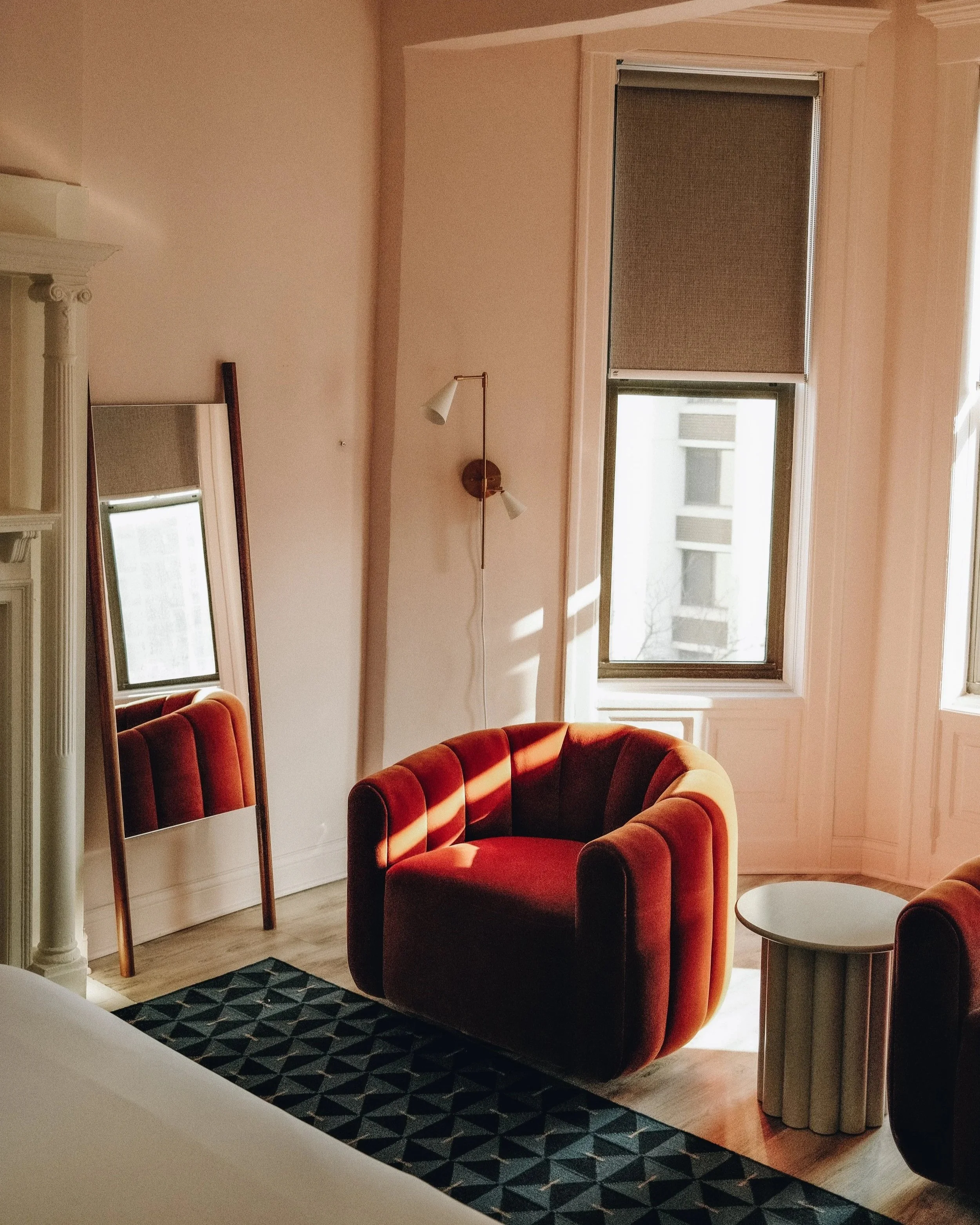 Living room with a red velvet armchair, a round white side table, sunlight coming through the window, a tall mirror leaning against the wall, and a wall-mounted light fixture.