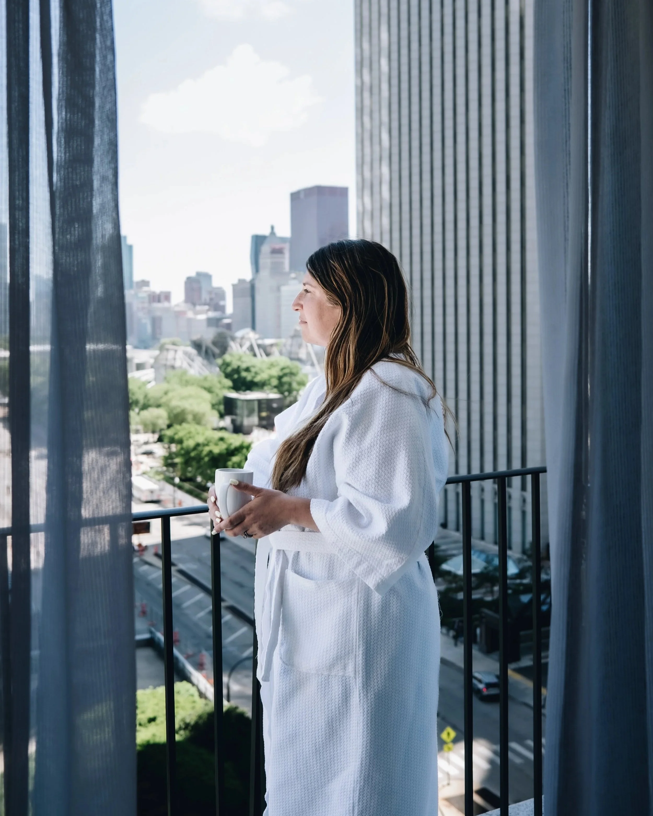 Woman standing on a balcony holding a white mug, with tall city buildings and a clear sky in the background.