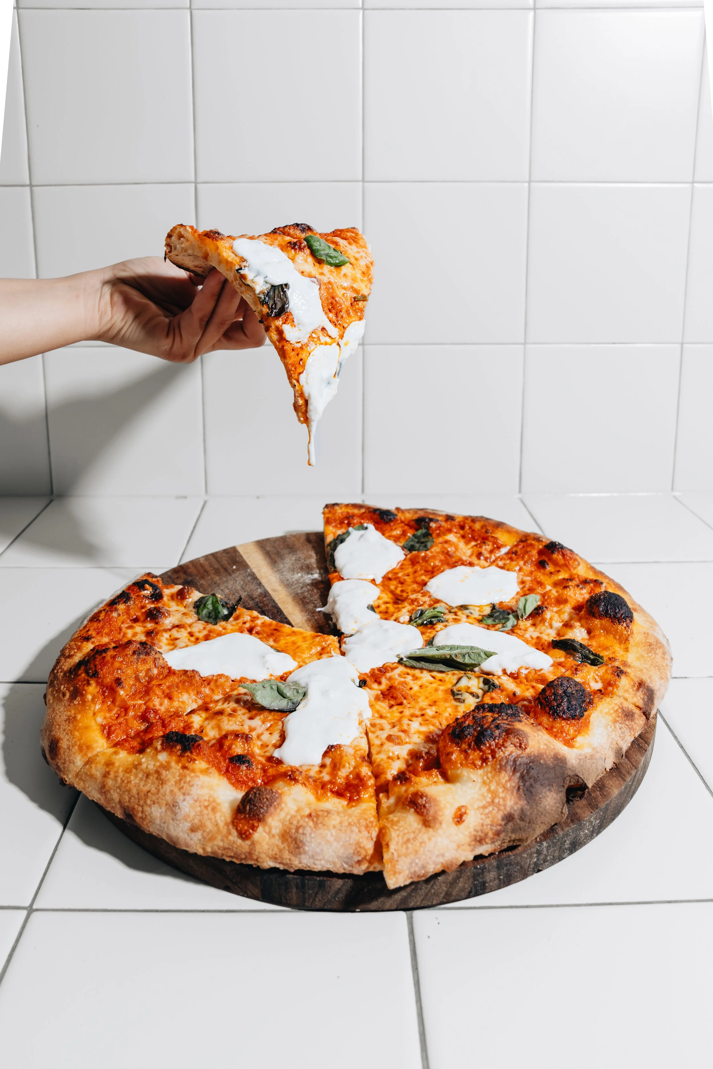 A person holding a slice of pizza with cheese, basil, and tomato sauce over a whole pizza with similar toppings on a wooden pizza board against white tiled wall.