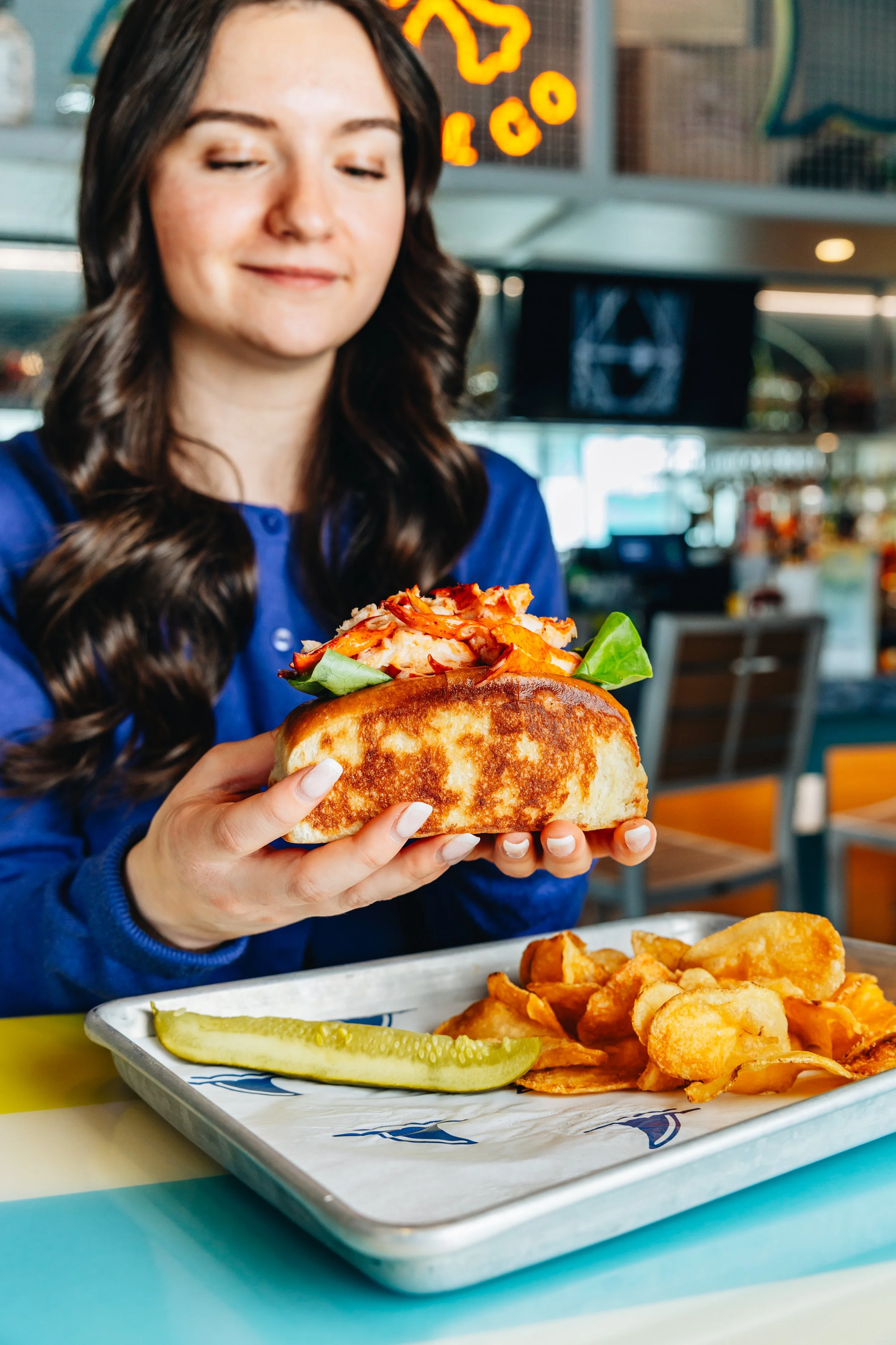 A woman in a blue shirt holding a sandwich with lettuce, chicken, and barbecue sauce in a restaurant. A tray with potato chips, fried pickles, and a pickle spear is on the table.