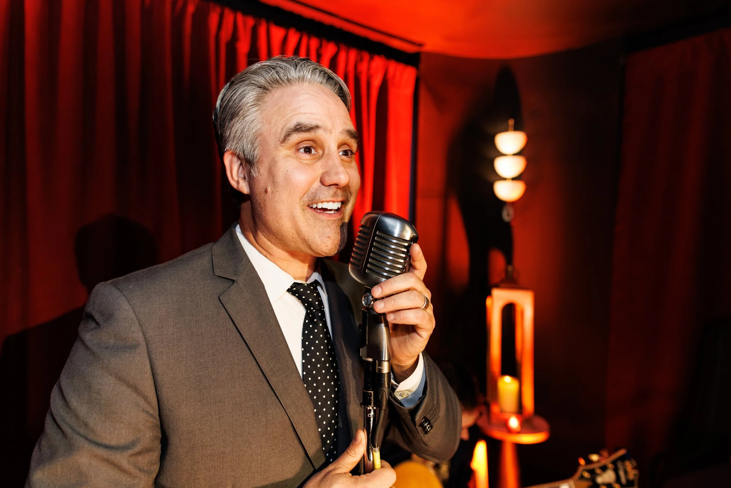 A man in a gray suit and polka dot tie singing into a vintage microphone in a dimly lit room with red curtains and warm lighting.