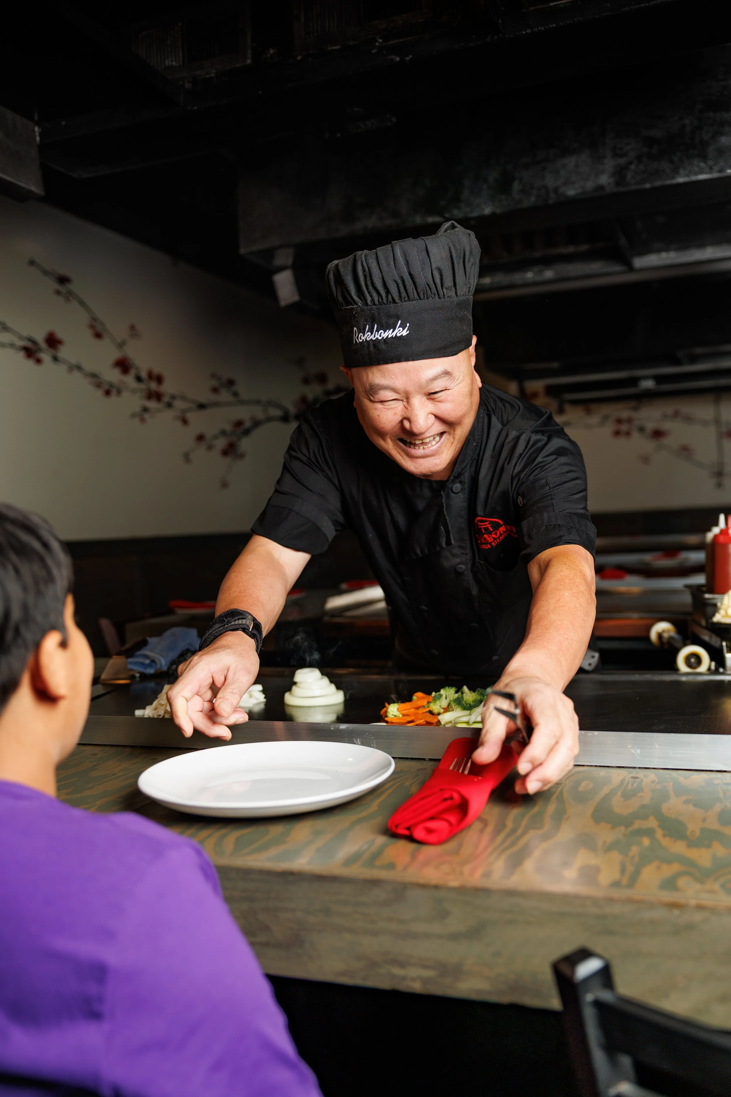A chef wearing a black hat with 'Rokkonki' embroidered on it and a black uniform is preparing food at a restaurant counter, with vegetables on a cutting board and a red napkin in front.