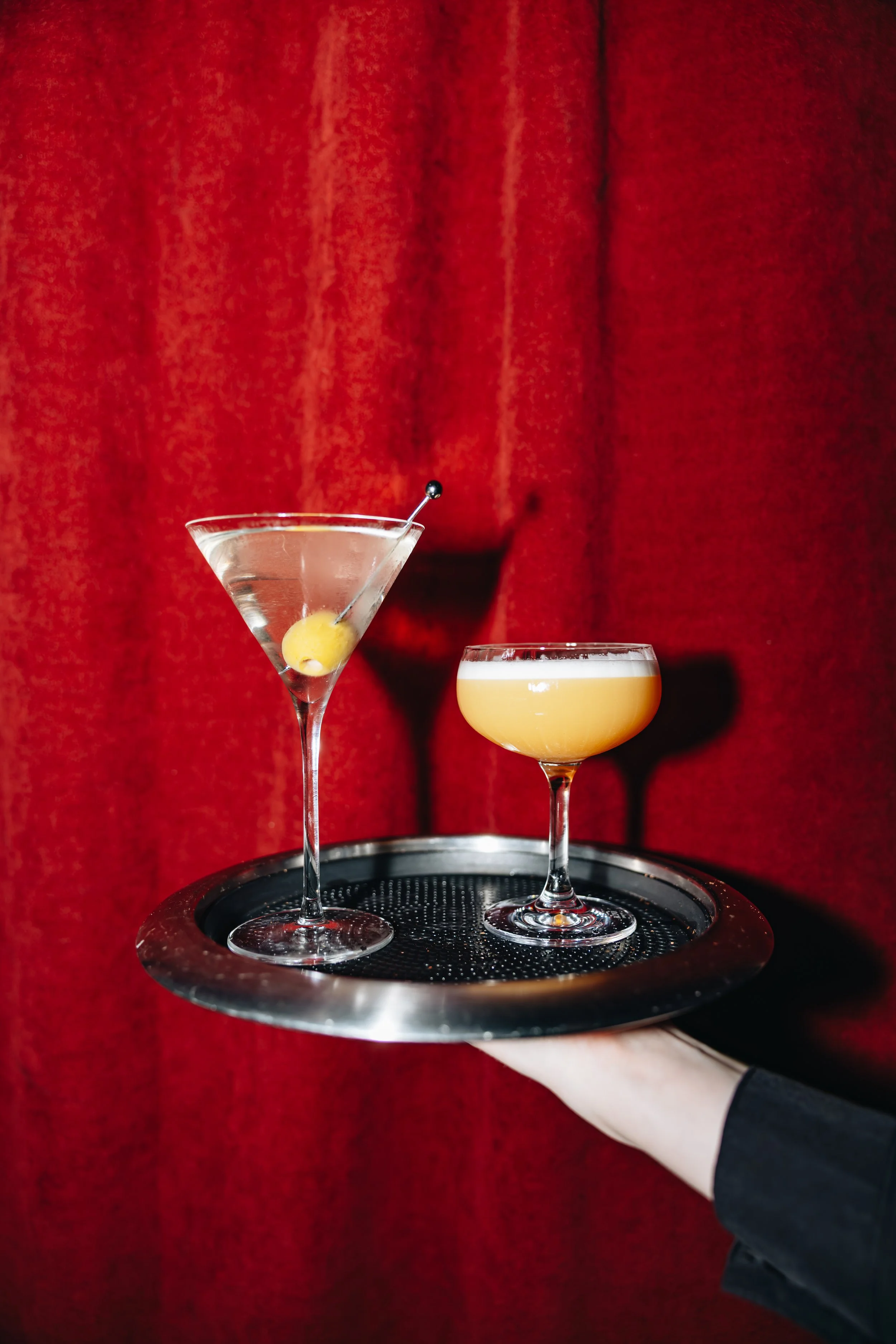 Hand holding a black tray with two different cocktails in front of a red curtain background.